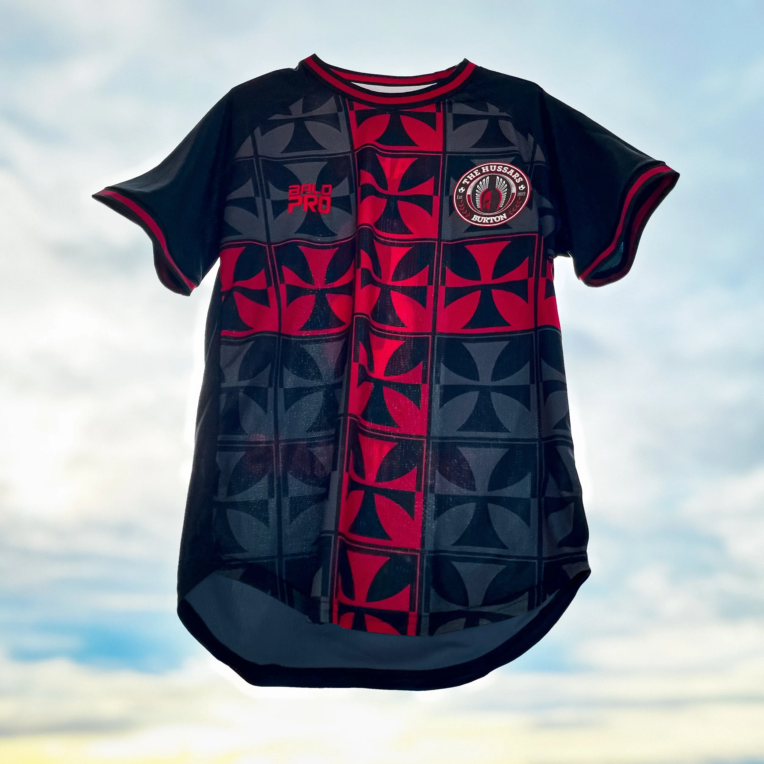 Ein dunkles Sportshirt mit rotem und schwarzem Muster, Logos und Text auf der Brust, gegen den Himmel fotografiert.