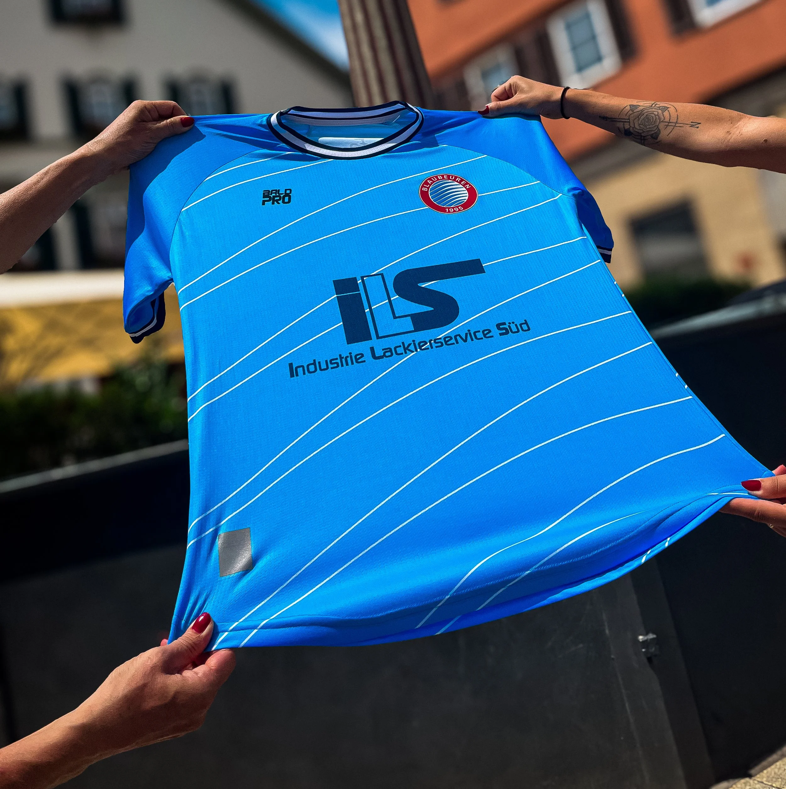 Zwei Personen halten ein blaules Trikot eines Fußballteams mit weißem Senkelmuster, einem Logo des Vereins Blau-Weiß Linnen, und Sponsorenlogo in der Mitte gegen die Kamera. Der Hintergrund ist eine Außenaufnahme mit Gebäuden.