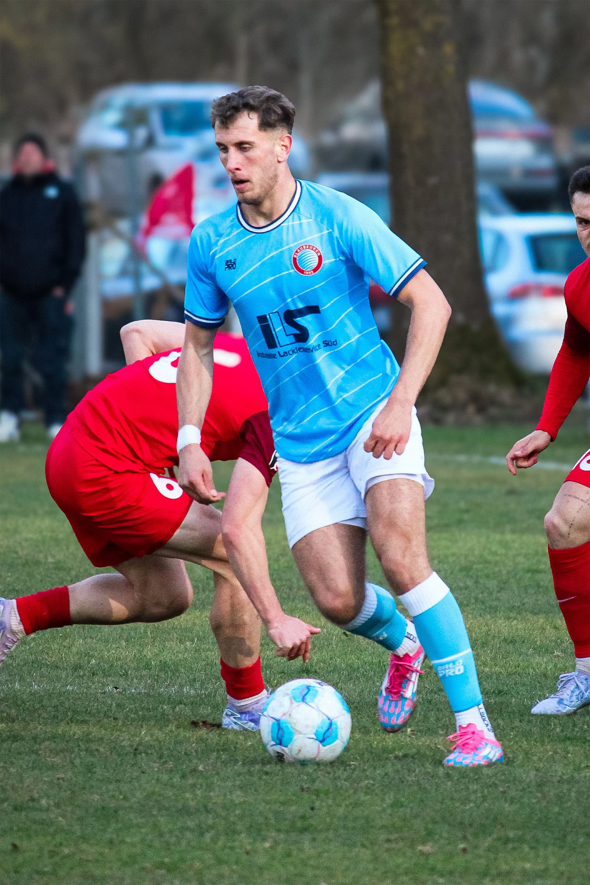 Fußballspieler in blauer Trikot und weißen Shorts dribbelt den Ball auf dem Spielfeld, während zwei Gegenspieler in roten Trikots versuchen, ihn zu stoppen.