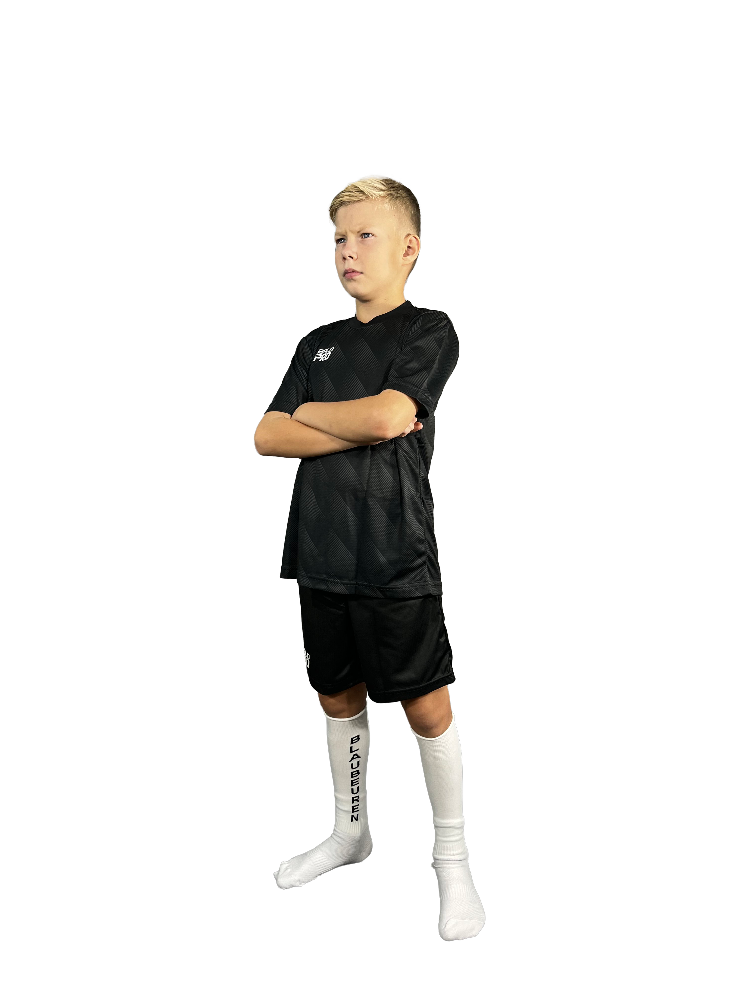 Ein junger männlicher Fußballspieler steht mit verschränkten Armen vor einem schwarzen Hintergrund und trägt ein schwarzes Fußballtrikot, weiße Socken mit schwarz geschriebenem Logo, und Fußballschuhe.