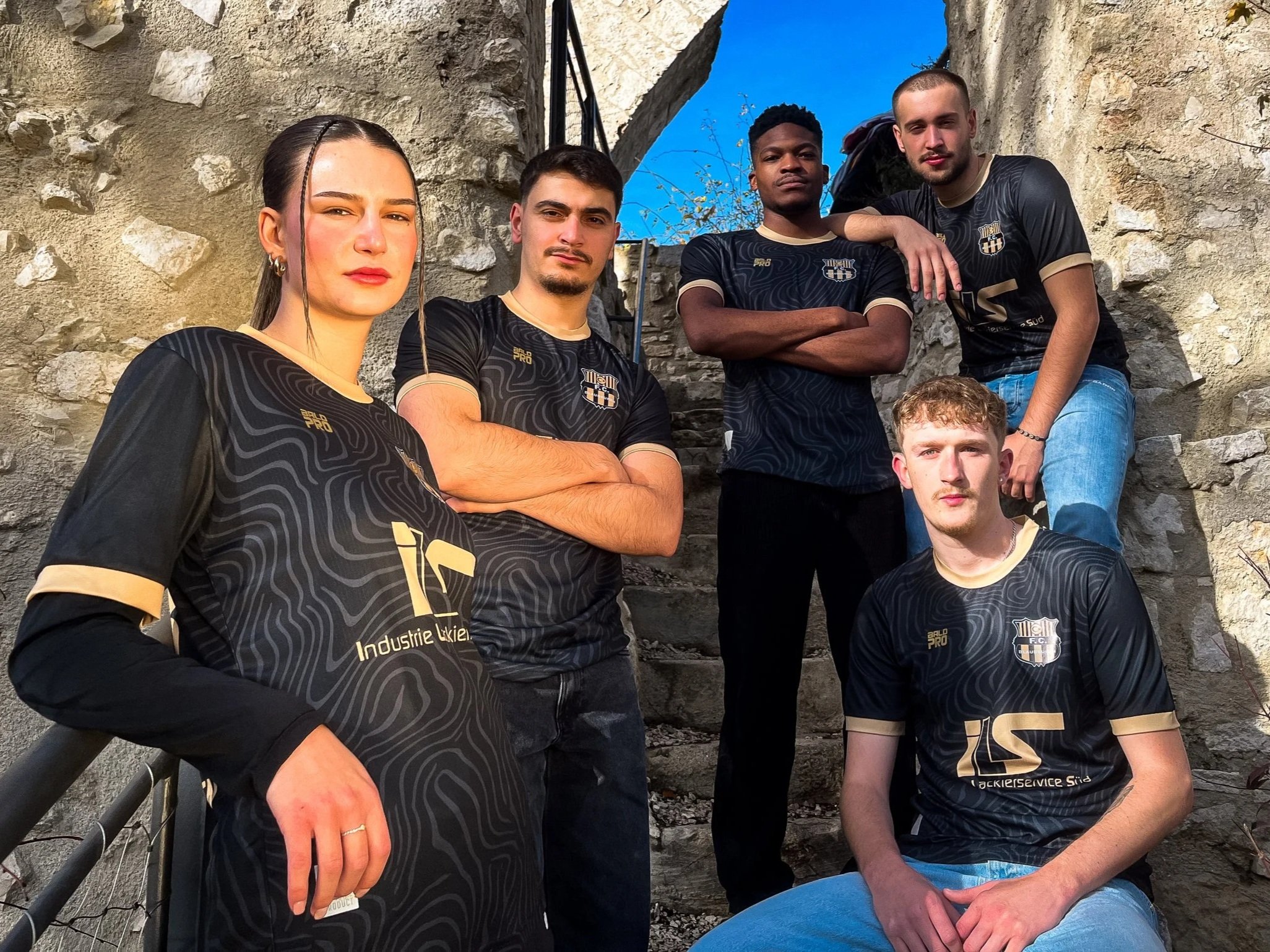 Sechs junge Menschen tragen schwarze SportTrikots und posieren auf einer Steintreppe vor einer alten Steinmauer. Es ist sonnig und der Himmel ist blau.