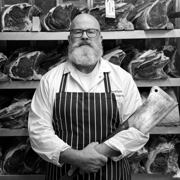 A man with glasses and a beard, wearing a striped apron and white shirt, holding a meat cleaver in front of a background of large cuts of meat on shelves.