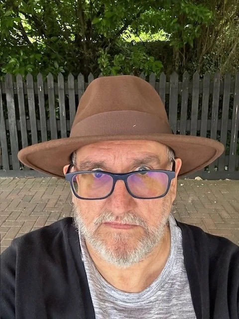 A man with glasses and a beard taking a selfie outdoors, wearing a brown hat and a gray shirt.