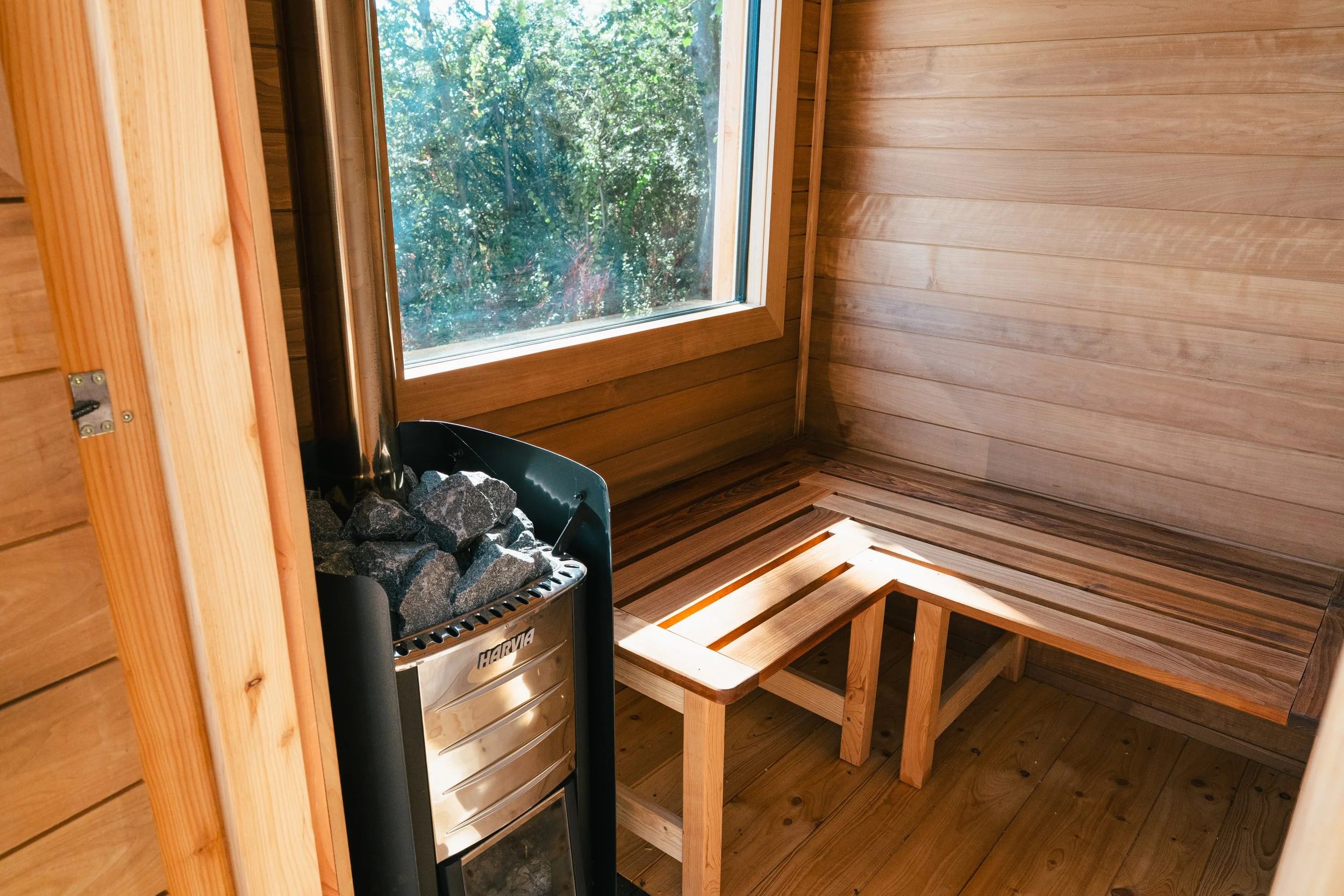 Interior of a wooden sauna room with benches, a window showing green trees outside, and a heater with sauna stones.