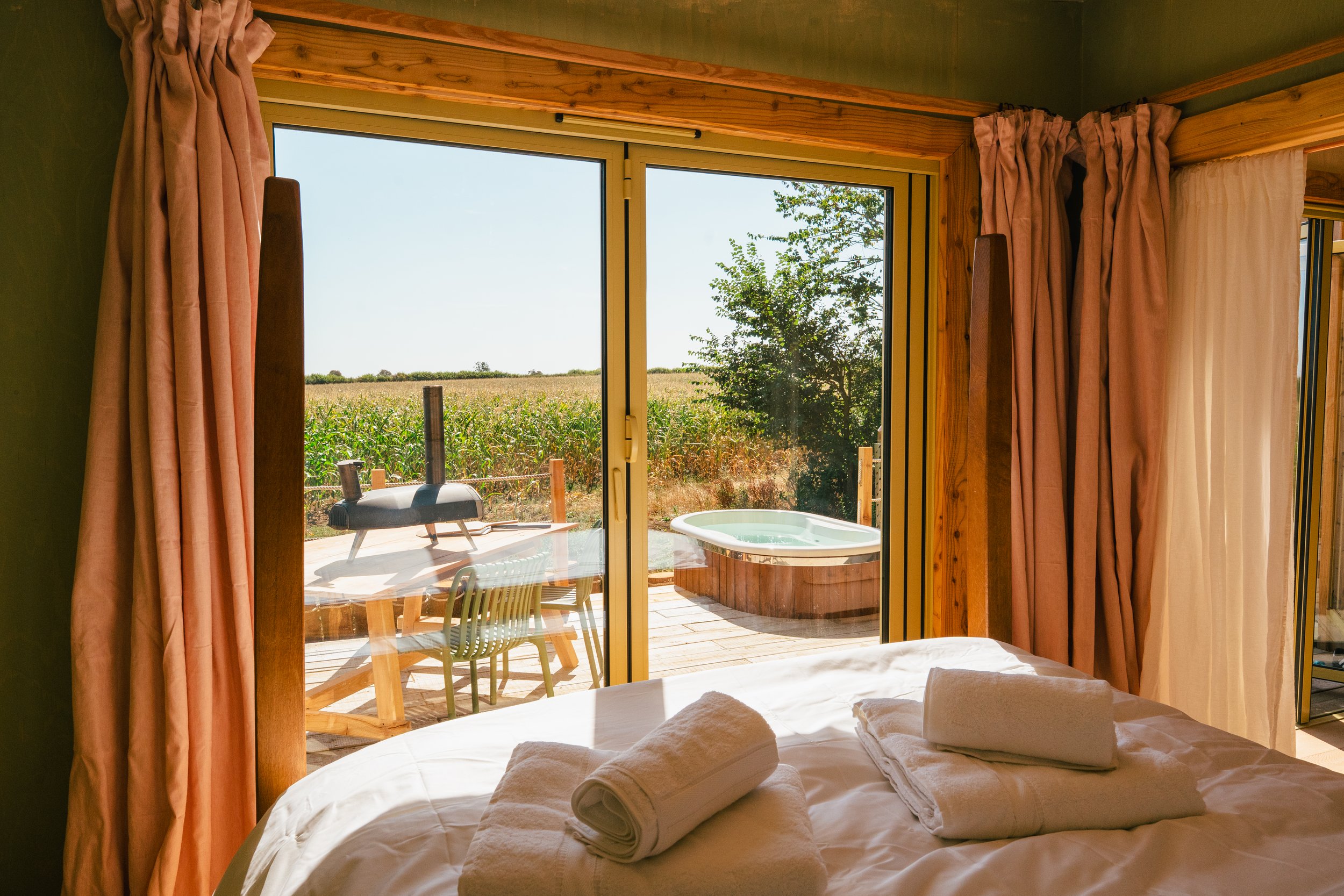 A bedroom view through sliding glass doors shows a deck with outdoor furniture, a hot tub, and a backyard with a field of crops and trees in the distance.