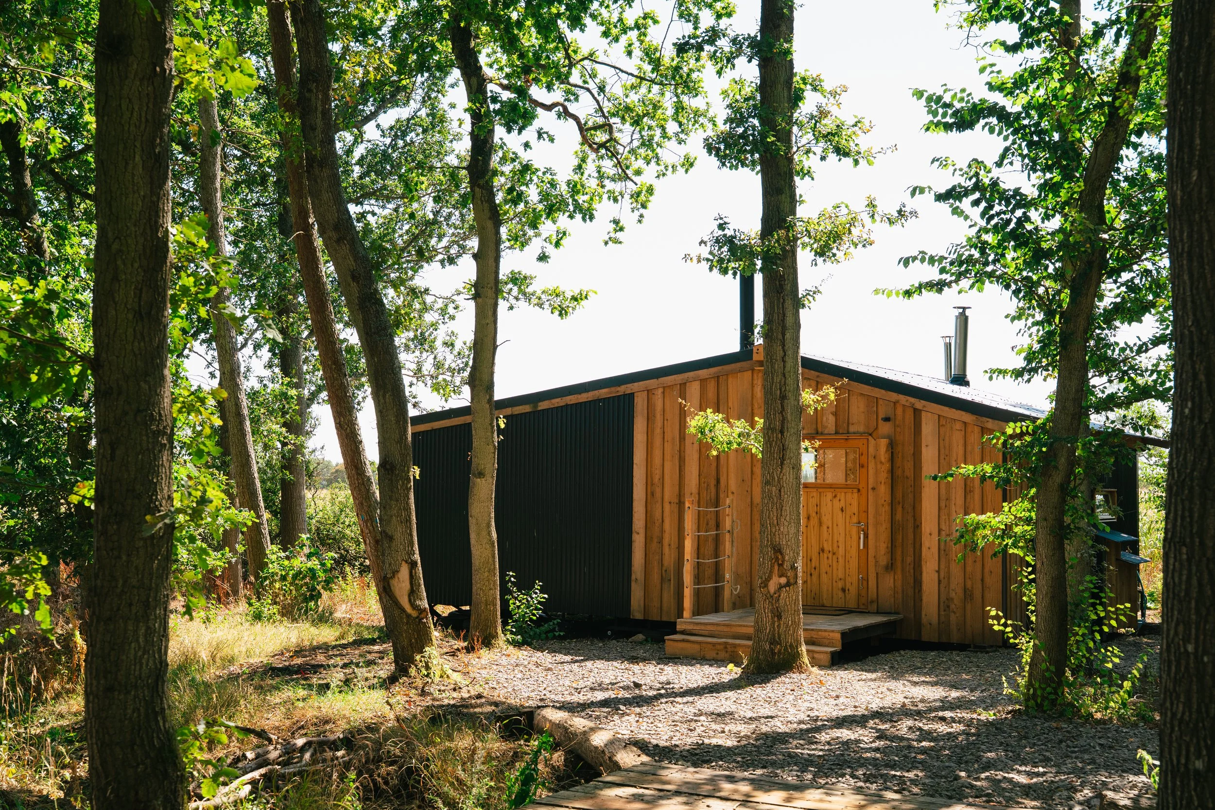 A small wooden cabin surrounded by trees with autumn-colored leaves on a cloudy day. All season woodland cabin in Cotswolds with hot tub near Cirencester and Bibury Village.