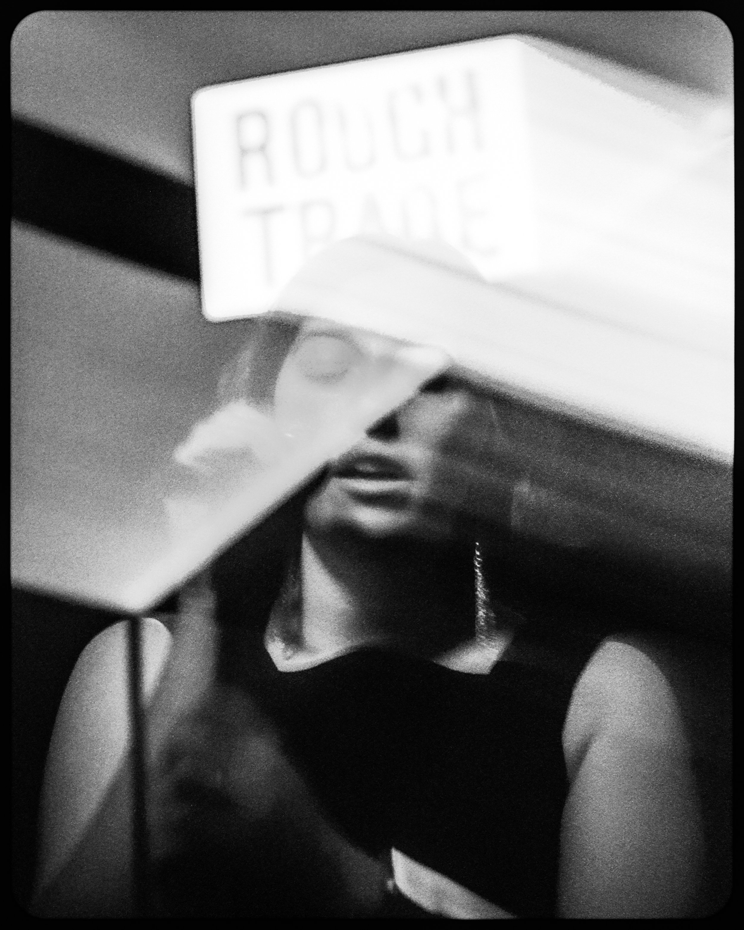 Black and white photo of a woman with glasses, partially obscured by a reflective surface, with a sign that says 'ROUGH TRADE' above her
