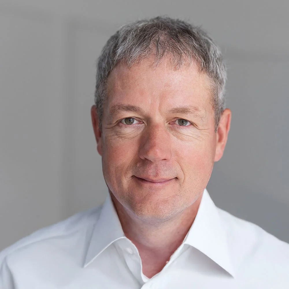 Portrait of Simon Edwards, a professional man with short gray hair, wearing a light blue checkered shirt, looking directly at the camera with a neutral expression.