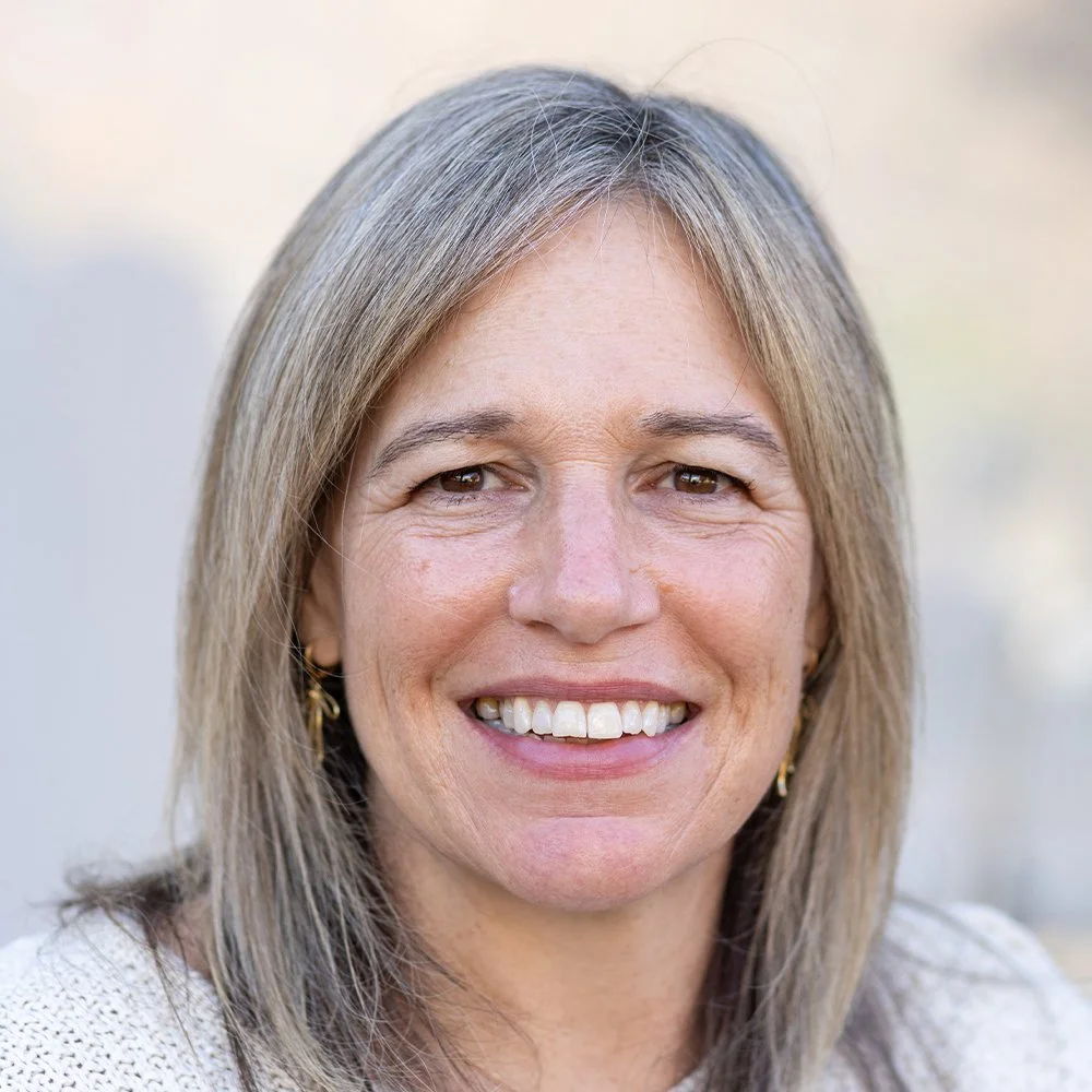 Headshot of Sara Cremins, a woman with shoulder-length ash blonde hair, wearing a textured white sweater, smiling at the camera with a warm expression.