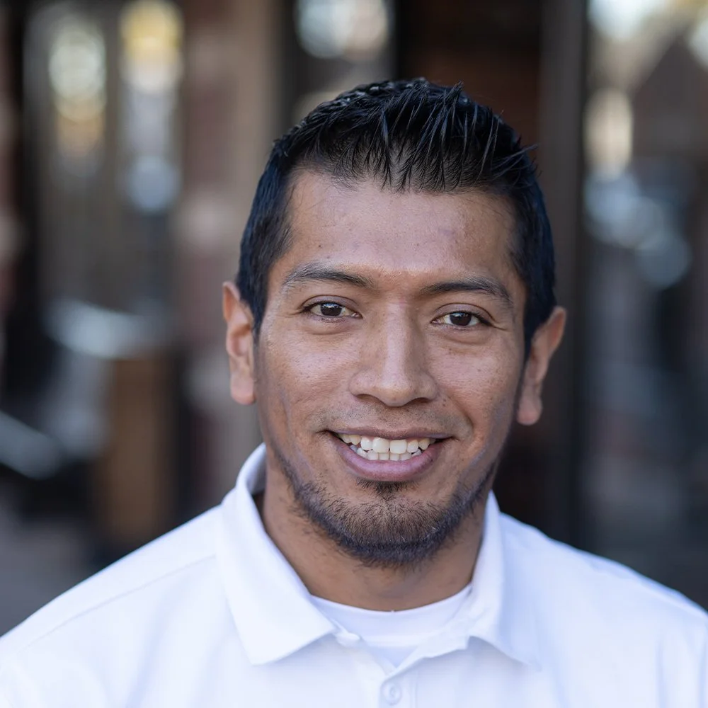 Portrait of Cristian Mayaguari, a man with short black hair, wearing a white polo shirt with a Hanley Group logo, smiling warmly at the camera.