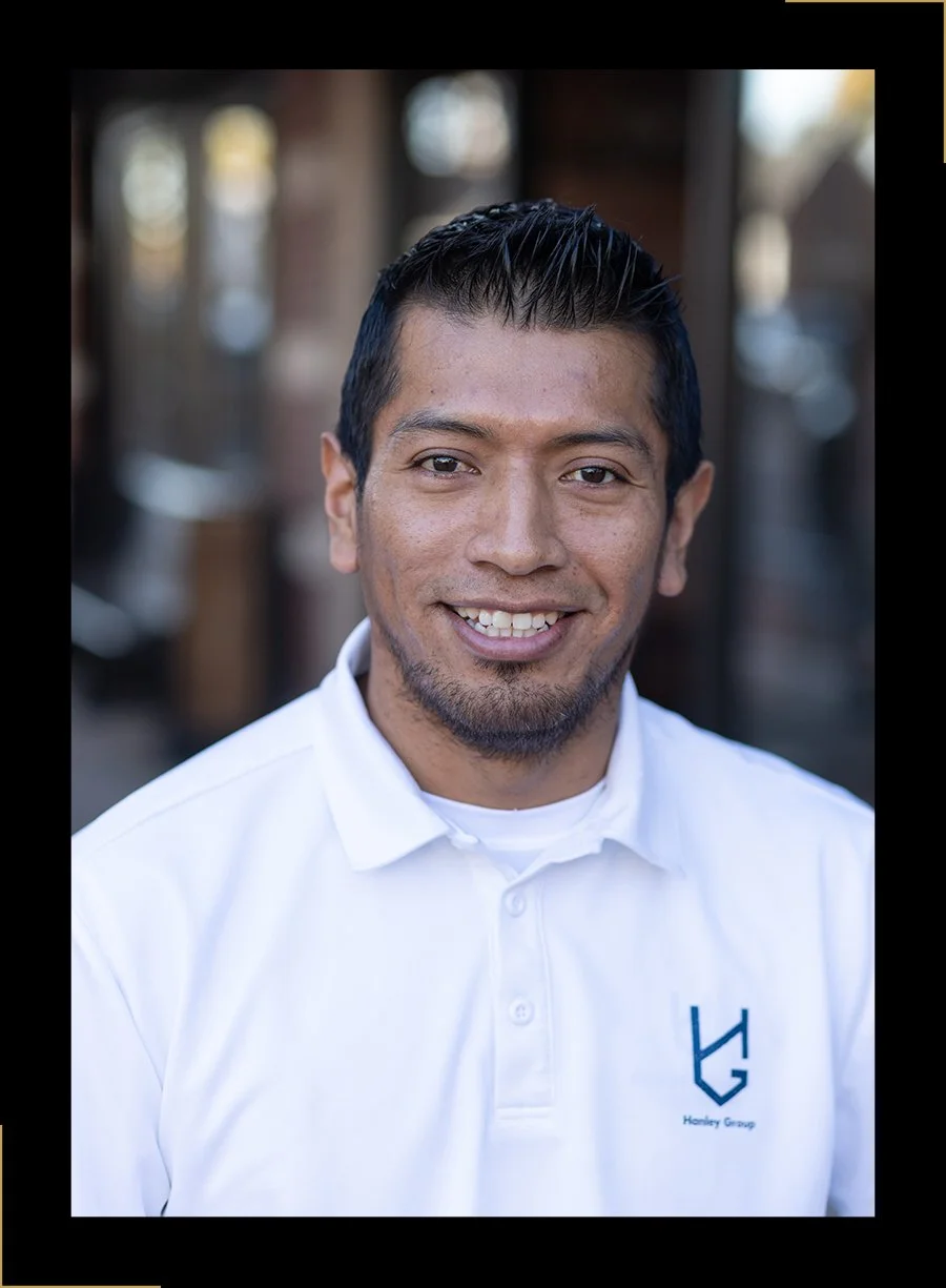 Portrait of Christian Mayaguri, a man with short black hair, wearing a white polo shirt with a Hanley Group logo, smiling warmly at the camera.
