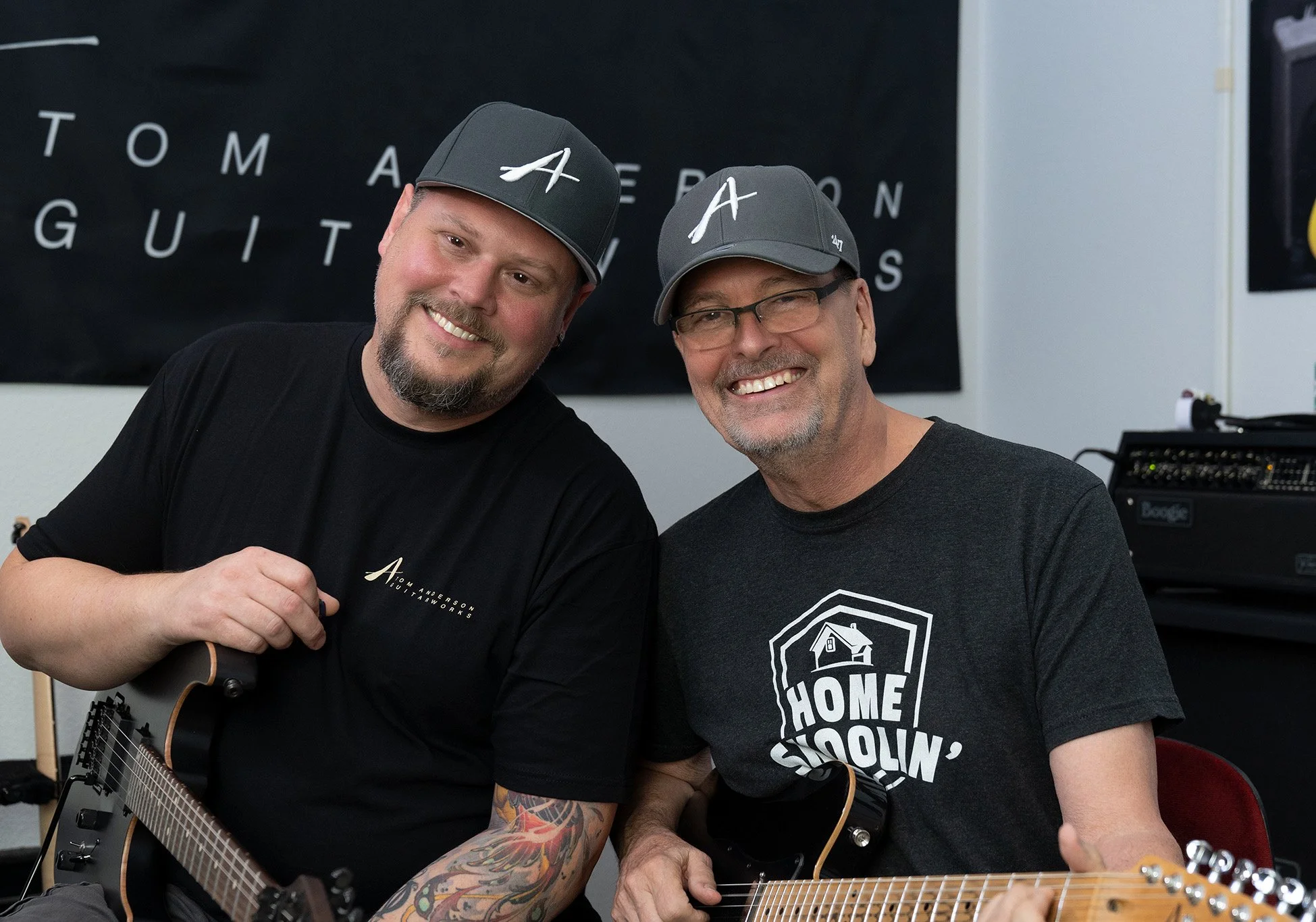 Markus Spohn and Tom Anderson smiling men wearing gray baseball caps and black t-shirts, one holding a guitar, sitting together in a music studio with equipment and a black banner that reads 'TOM ANDERSON GUITARSWORKS' in the background.