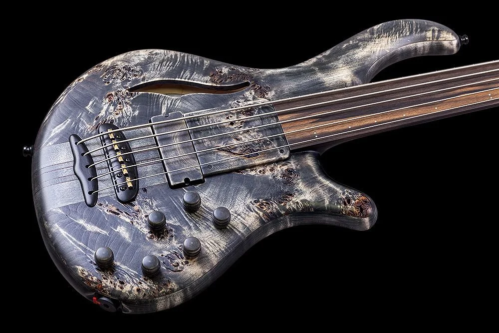 A close-up of a modern Mayones electric bass guitar with a unique wood grain finish, set against a black background.