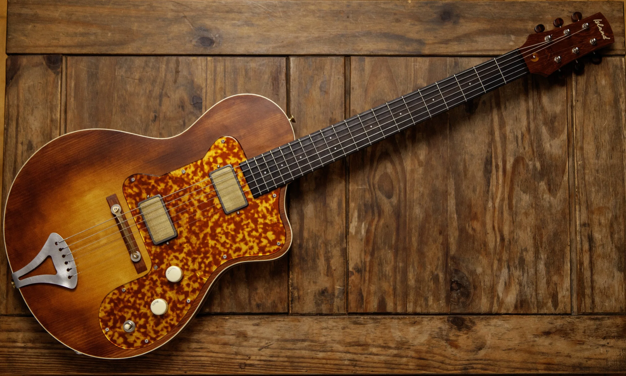 An electric Island Instruments guitar with a sunburst finish, tortoise shell pickguard, and vintage-style hardware, placed on a wooden floor.