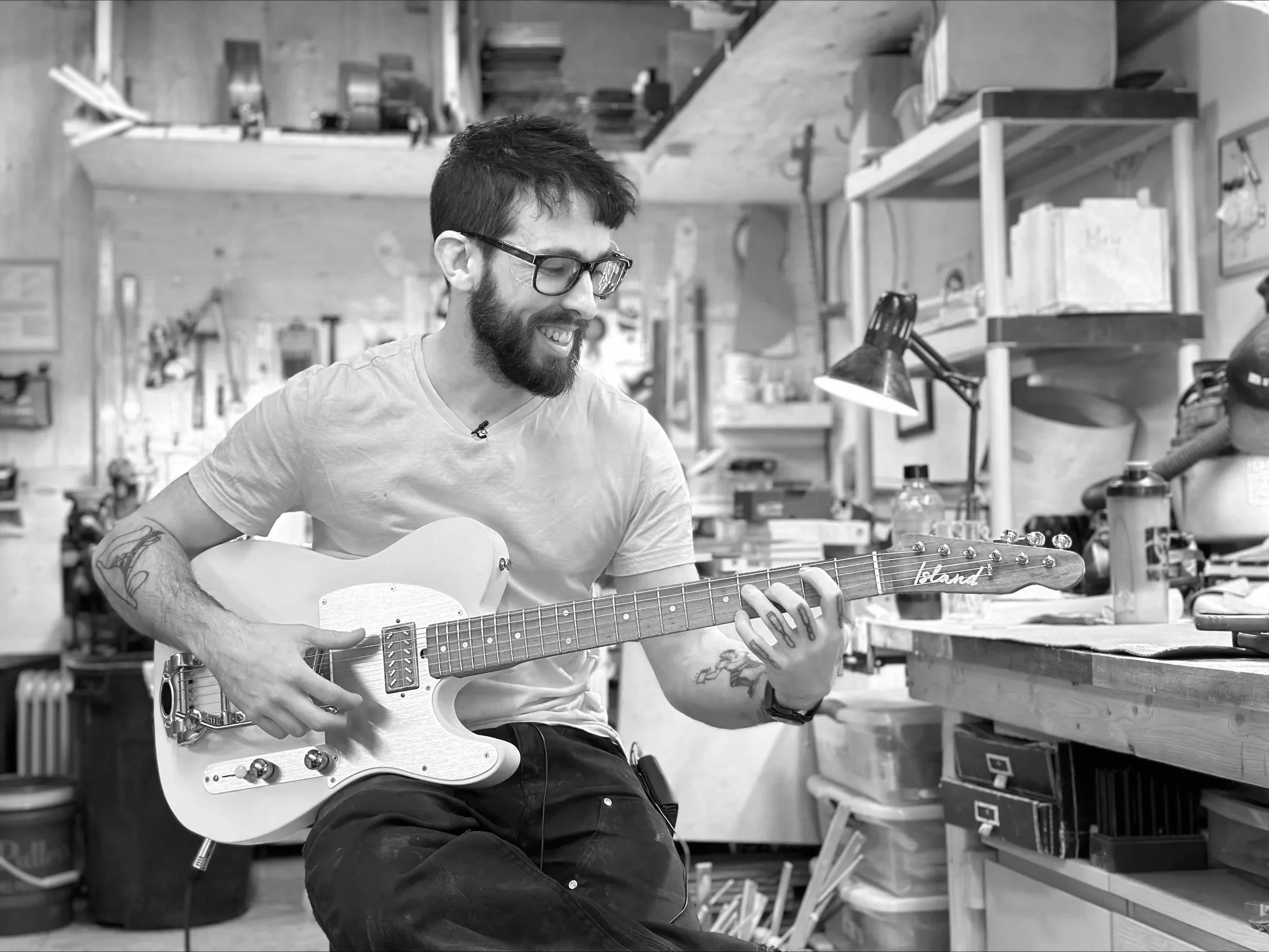 Nic Delisle with glasses and a beard playing an electric guitar in a workshop or music studio with shelves and tools in the background.