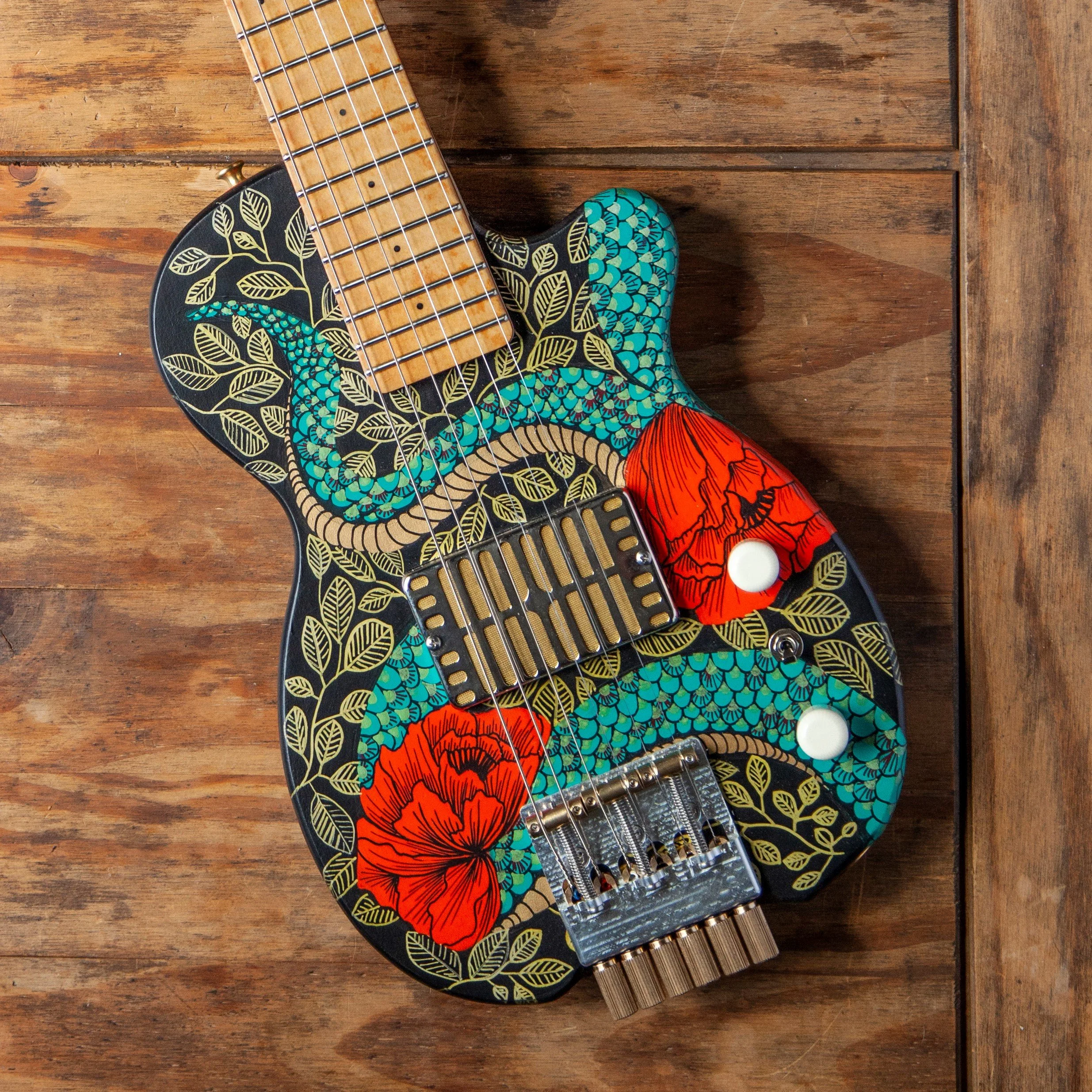 Close-up of a decorated electric Island Instruments guitar with black background, gold leaf and floral designs, resting on a wooden surface with a small karaoke microphone next to it.