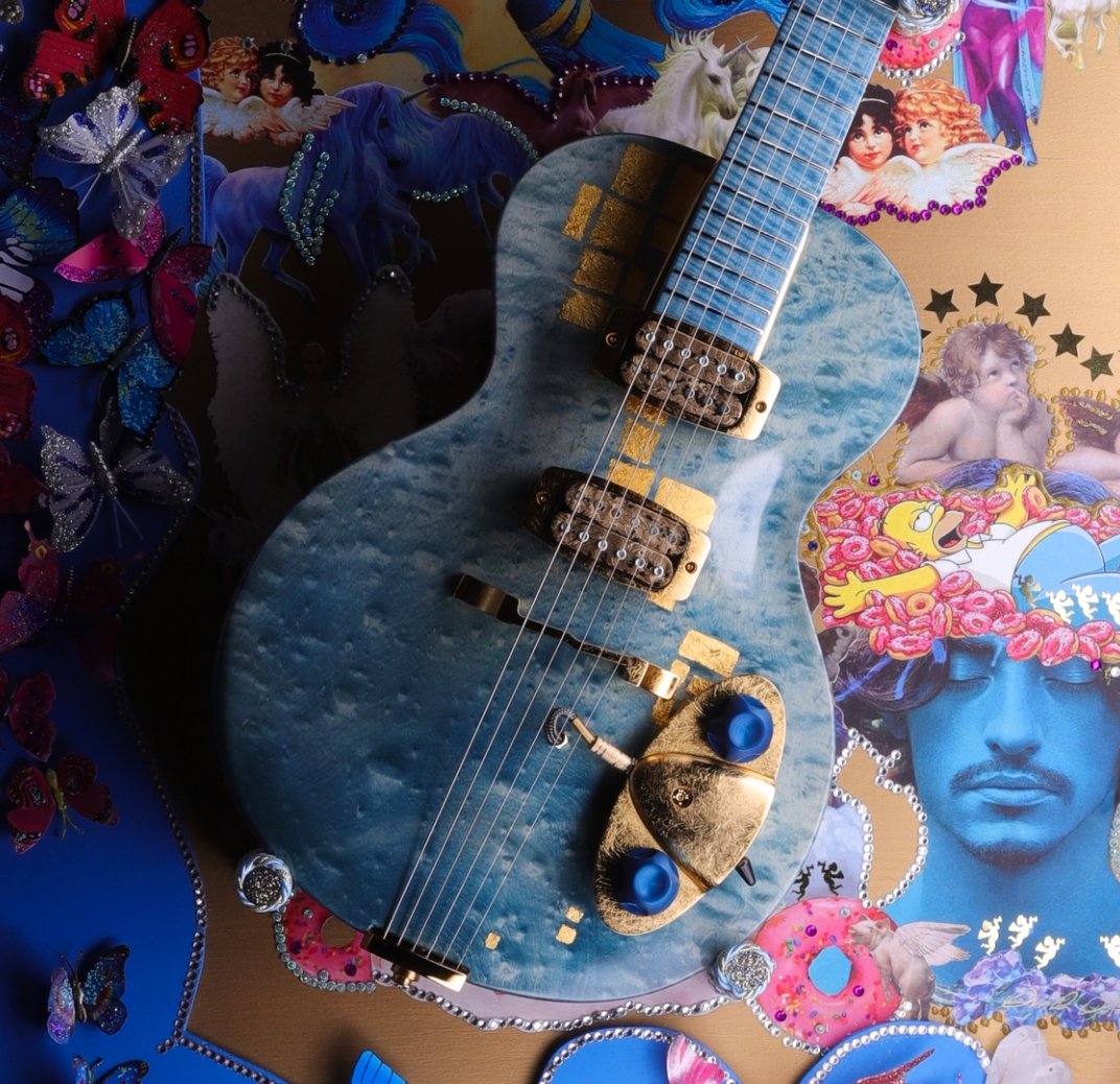 A blue electric guitar with gold accents placed on a colorful collage background featuring butterflies, angels, and vibrant art.