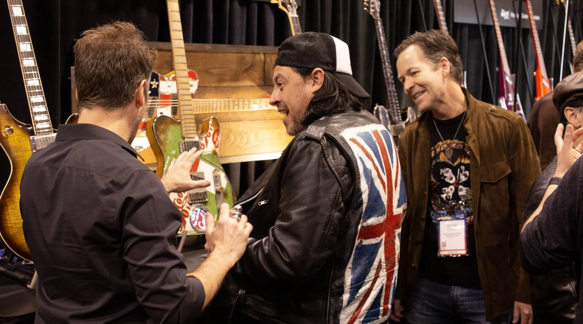 Men examining guitars on display at a music convention, one wearing a jacket with a Union Jack design.