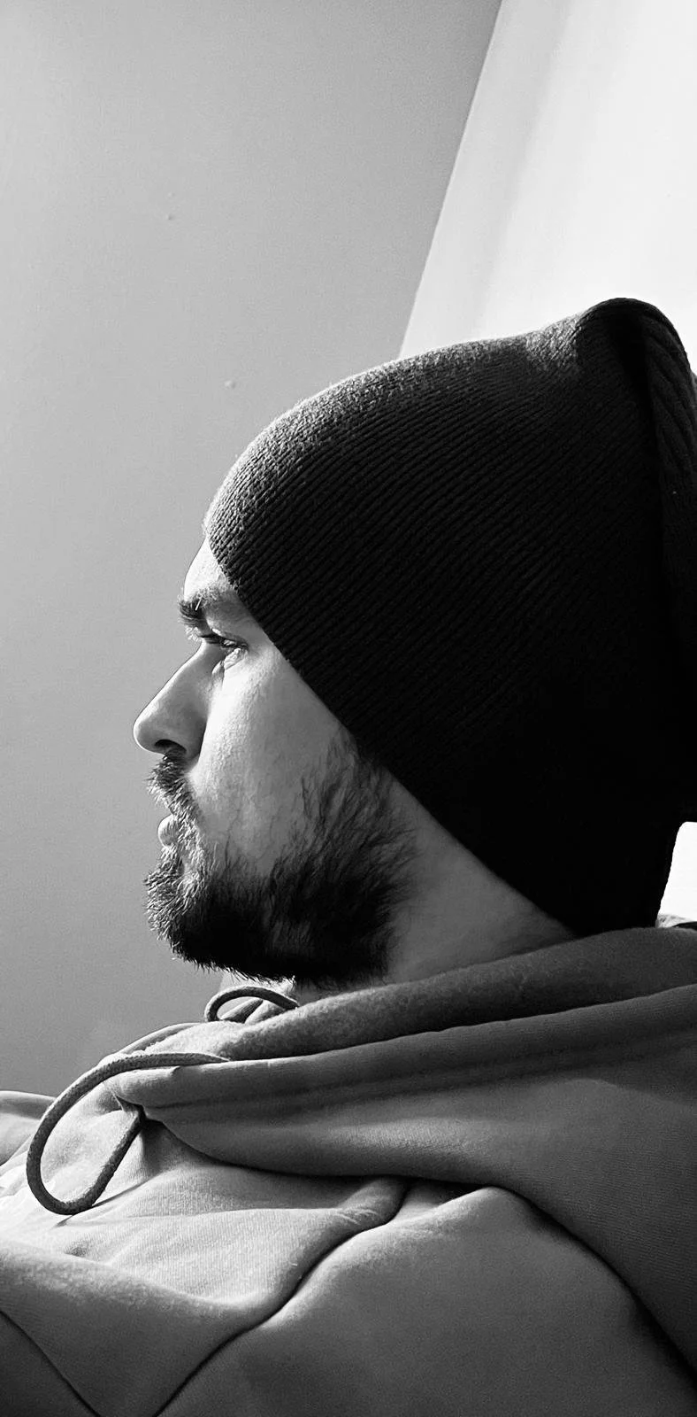 A black and white profile of Simon Padalka with a beard, wearing a beanie and a hoodie, sitting against a plain wall.