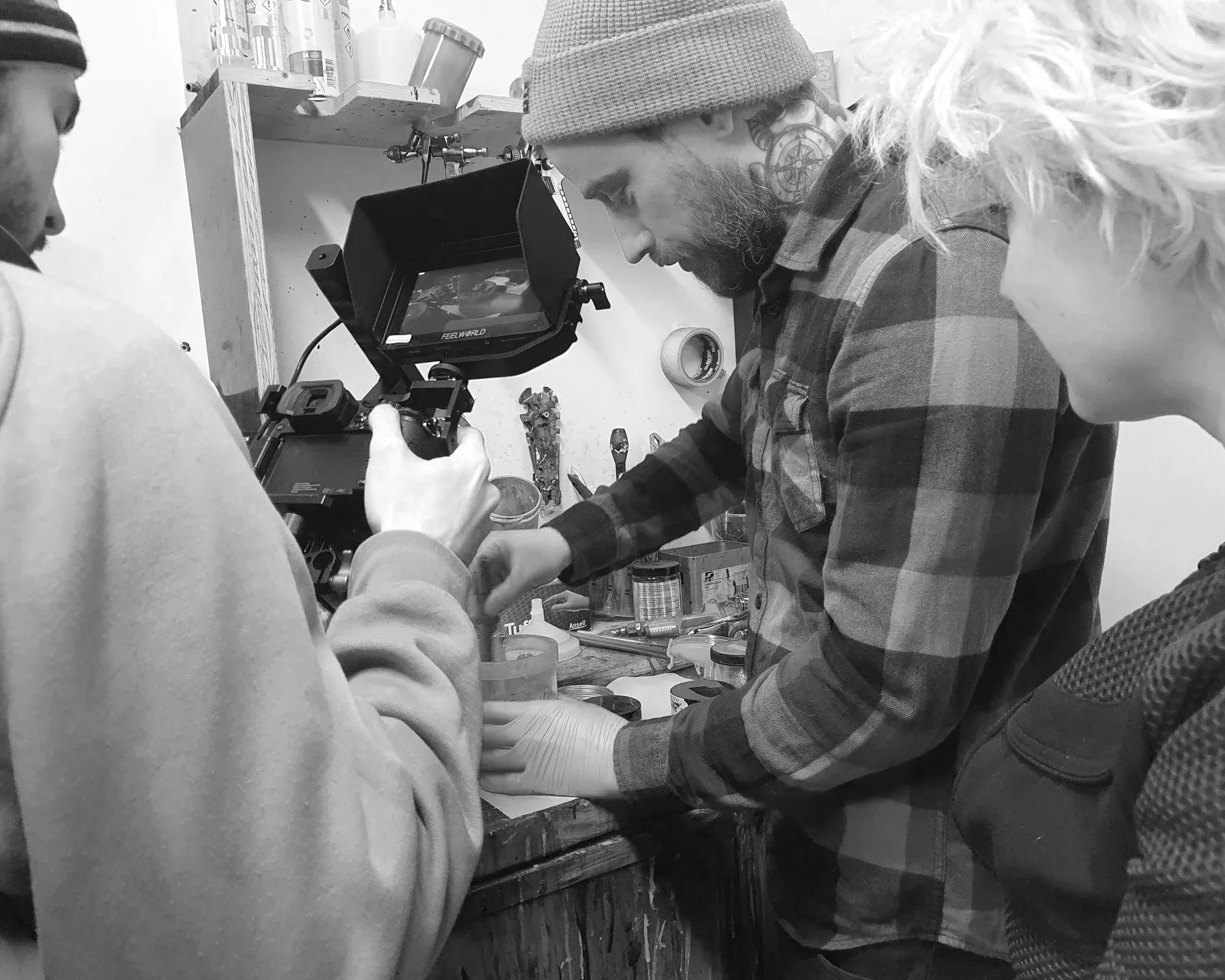 Three people working together in a workshop, with one person holding a camera to film the other two who are handling tools and small objects on a workbench.