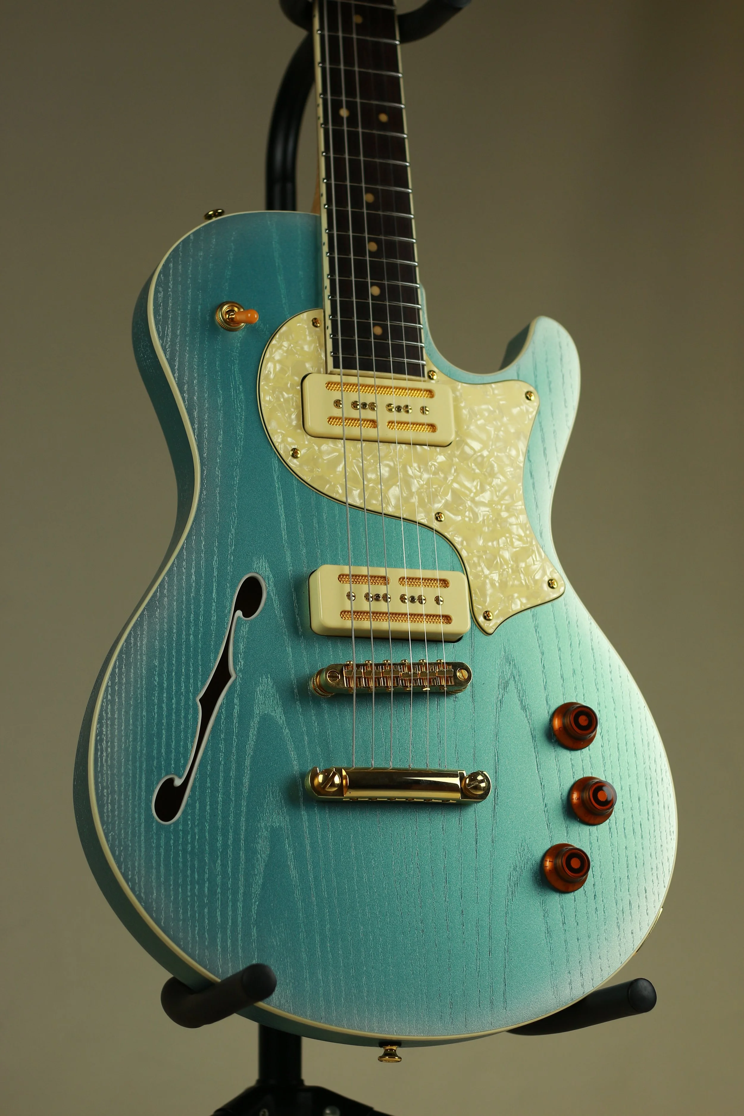 Blue semi-hollow Corsell electric guitar with white pickguard and orange control knobs on a black stand.