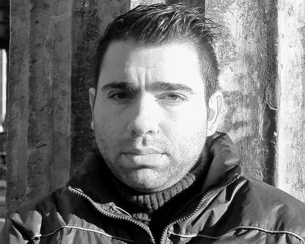 Alejandro Ramirez, a man with short dark hair and a serious expression, wearing a dark jacket and a black turtleneck, standing outdoors against a textured stone wall.