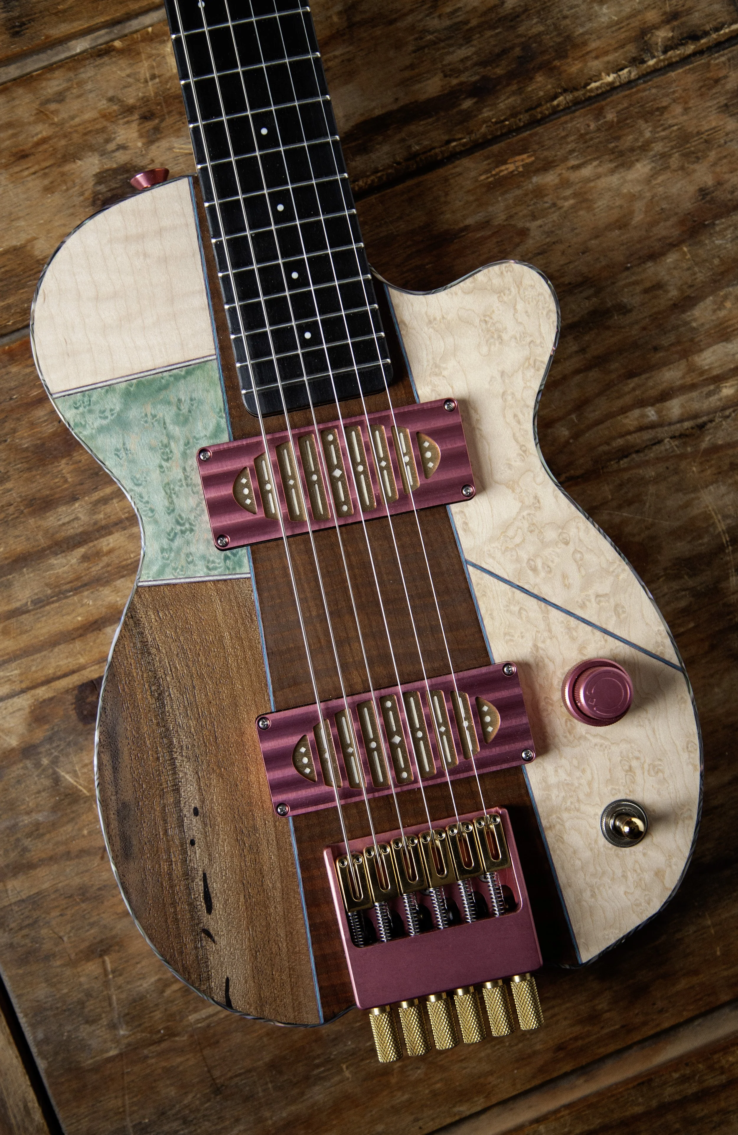 Close-up of an electric Island Instruments guitar with a multicolored body, black fretboard, unique pickups and hardware, resting on a wooden surface.
