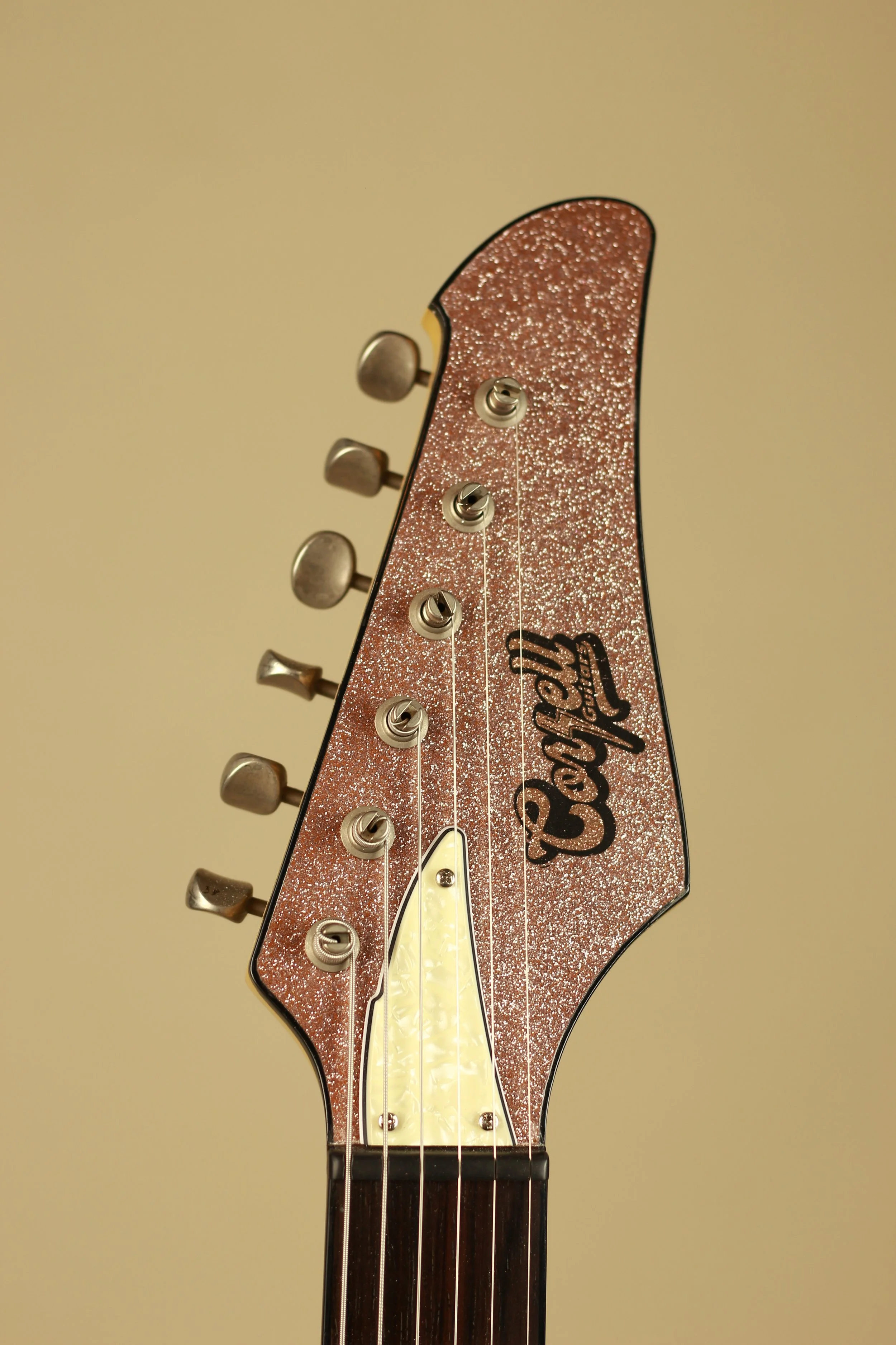 Close-up of a guitar headstock with a glittery pink finish, tuning pegs, and a yellow pearloid truss rod cover, and the word 'Corsell' written on it.