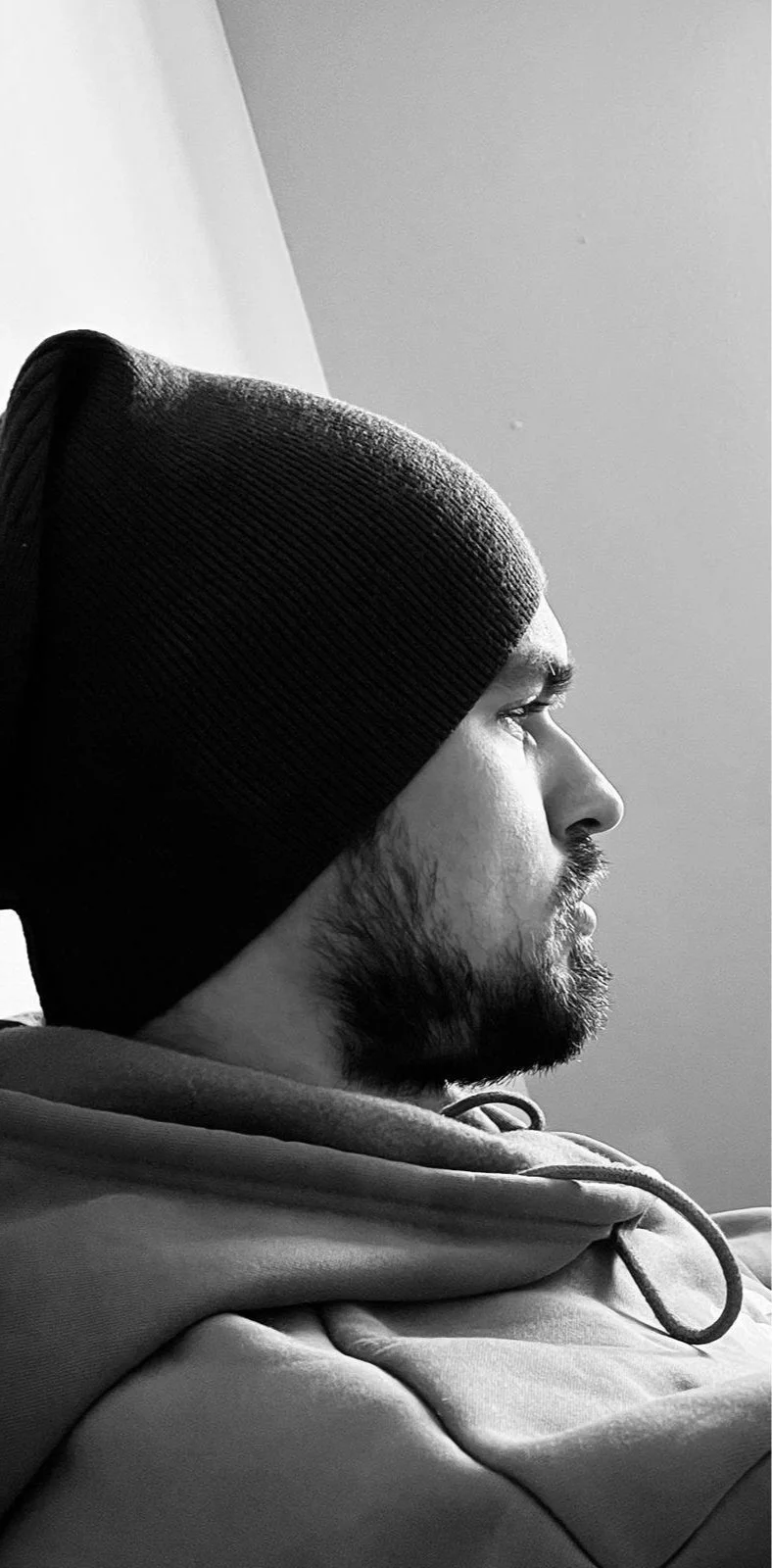 A black and white profile of Simon Padalka with a beard, wearing a beanie and a hoodie, sitting against a plain wall.