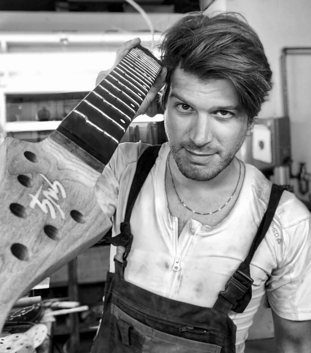Giulio Negrini, with styled hair and a slight beard, holding a guitar neck on his shoulder, smiling at the camera, in a workshop or studio setting.