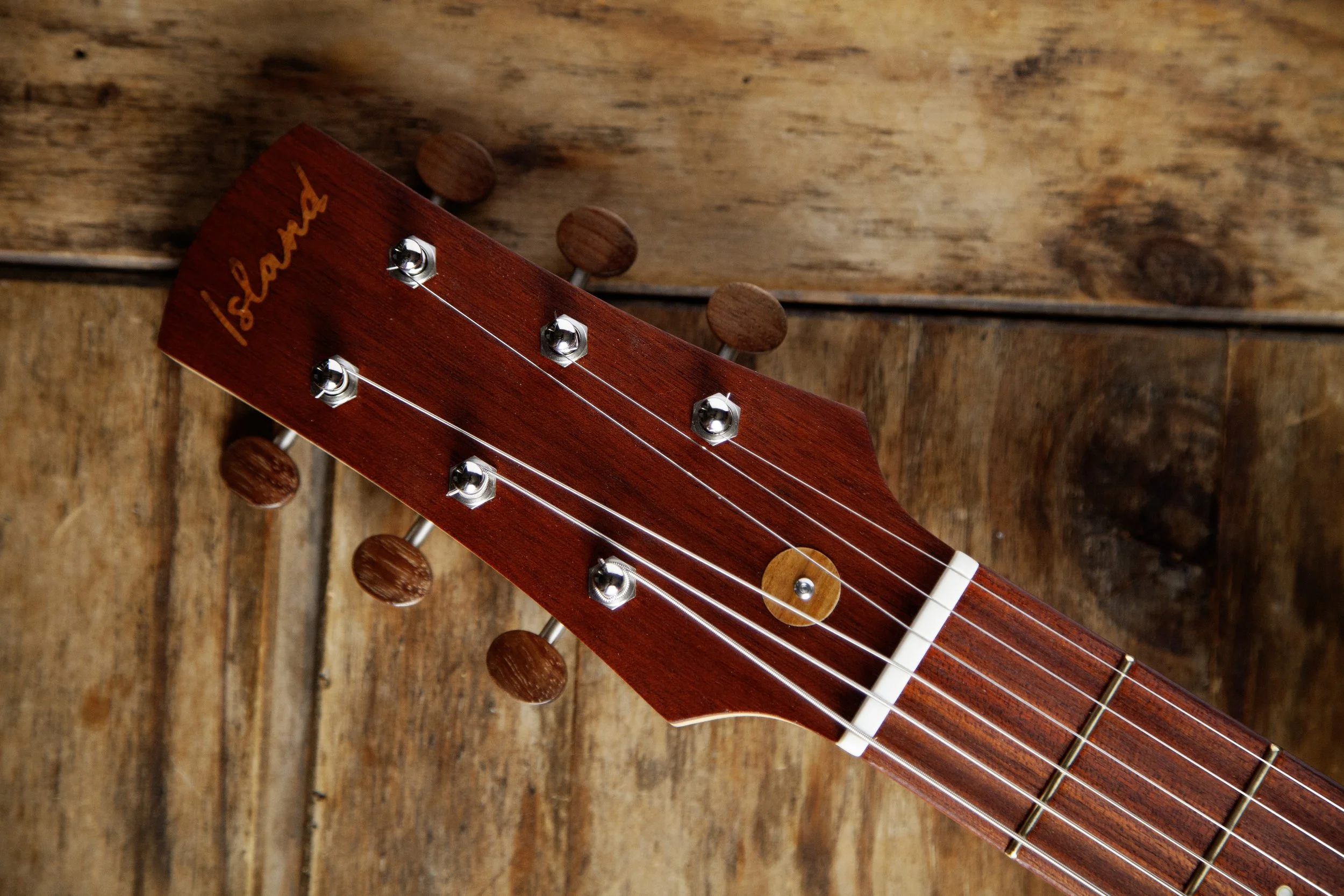 Close-up of a red wooden Island Instruments guitar headstock with tuning pegs and strings, resting on a wooden surface.