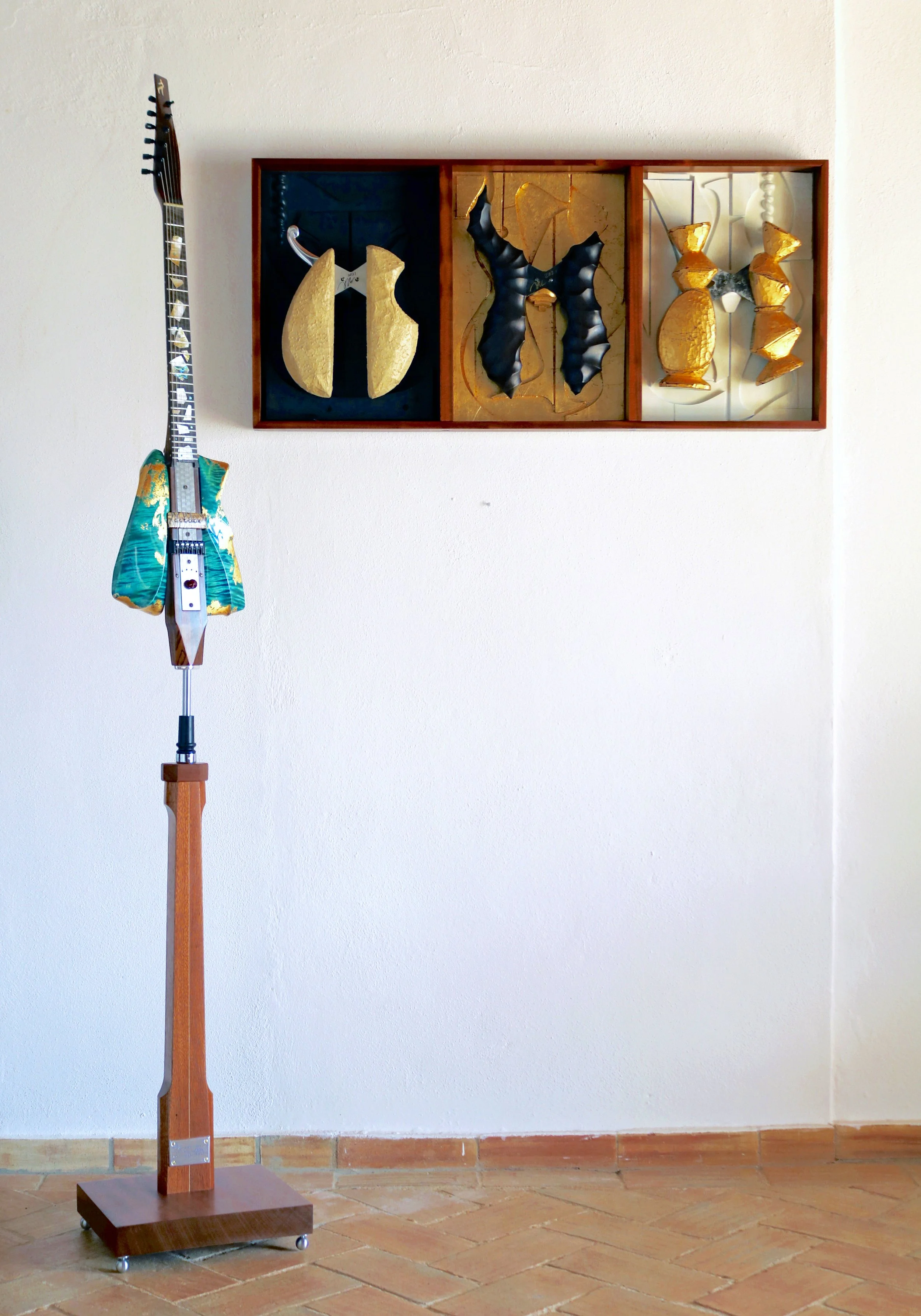 An art display featuring a mounted electric Spalt guitar with a colorful design and a wooden stand, next to a three-section wall art piece with abstract shapes in gold, black, and white.