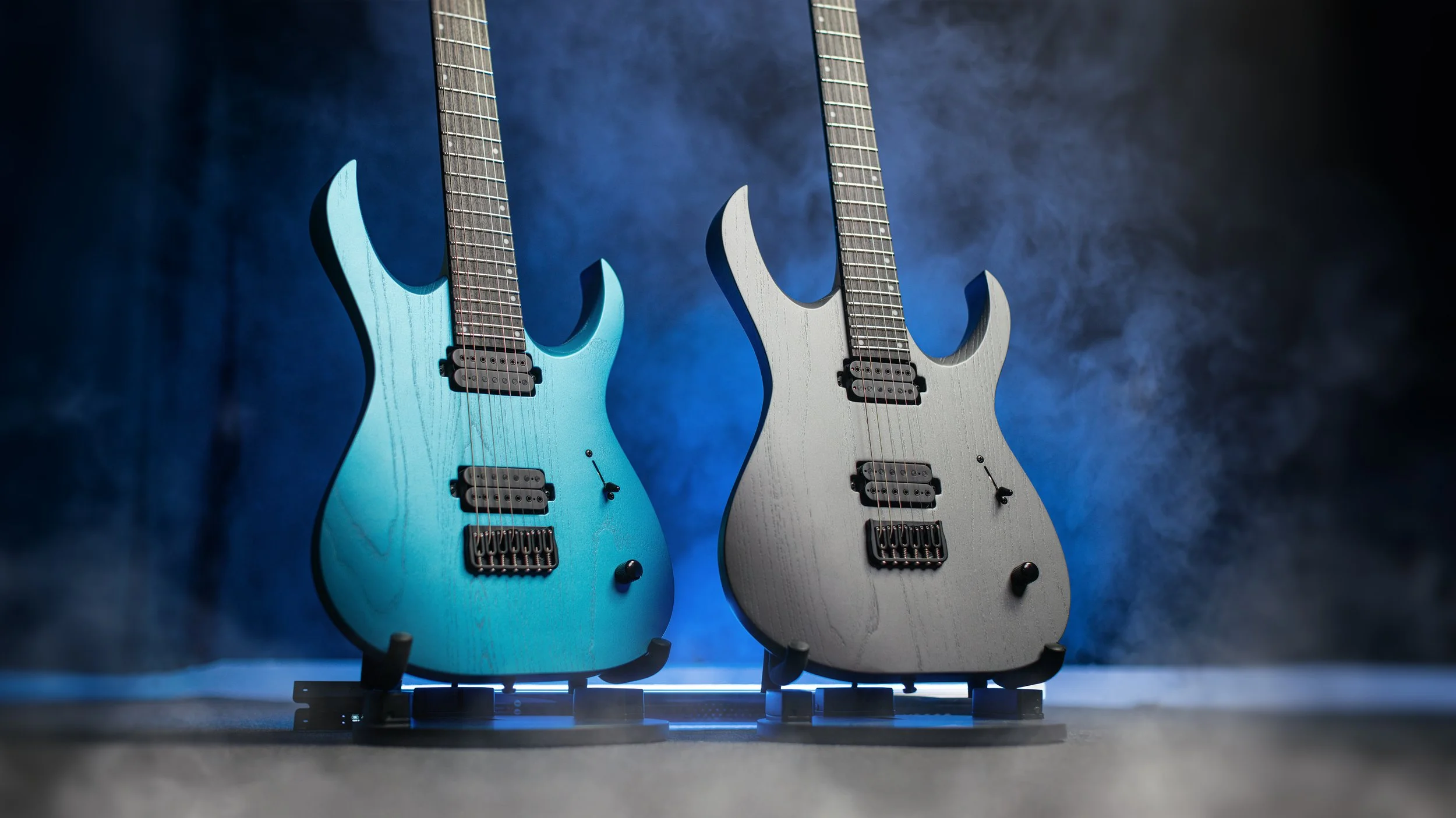 Two electric Mayones guitars, one blue and one gray, with pointed body shapes, black pickups, and knobs, standing on black stands against a dark, smoky background.