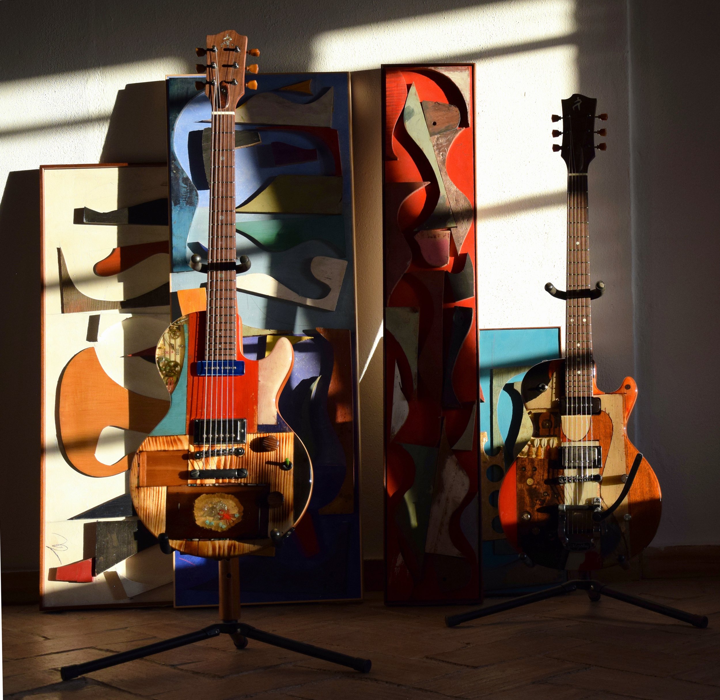 Two electric Spalt guitars are placed in front of abstract, colorful paintings on a wall, with sunlight casting shadows.