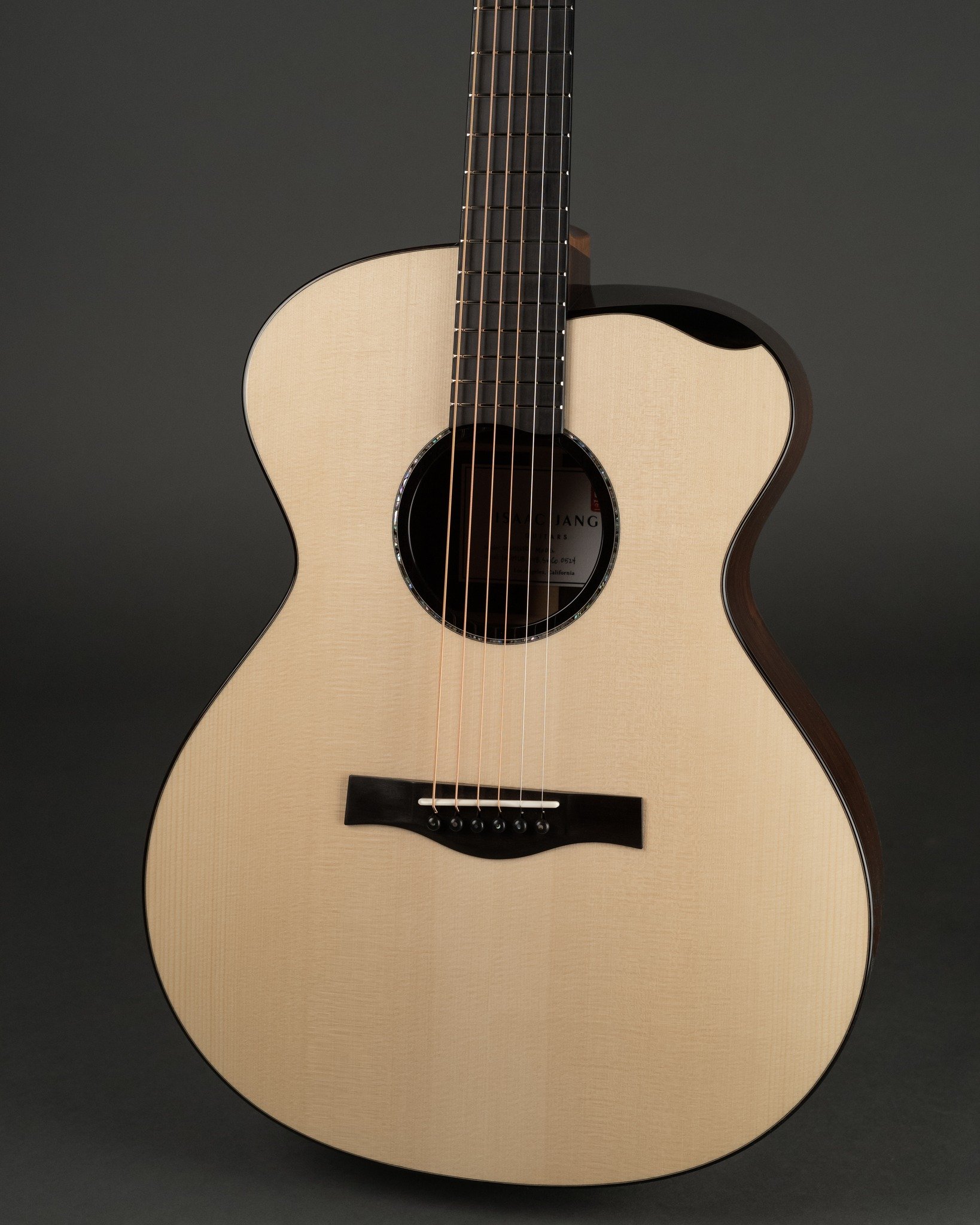 Isaac Jang Guitar close-up of an acoustic with a light-colored wooden body, black bridge, and dark fretboard, set against a dark background.