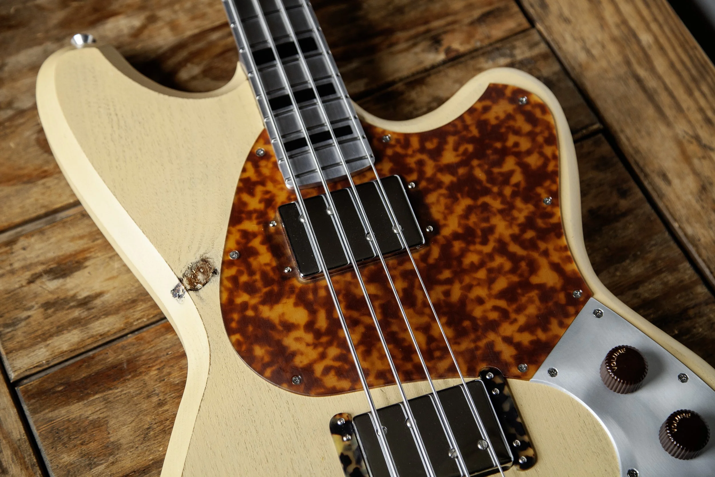 Close-up of an electric Island Instruments bass guitar with a tortoiseshell pickguard, metal strings, and control knobs, resting on a wooden surface.