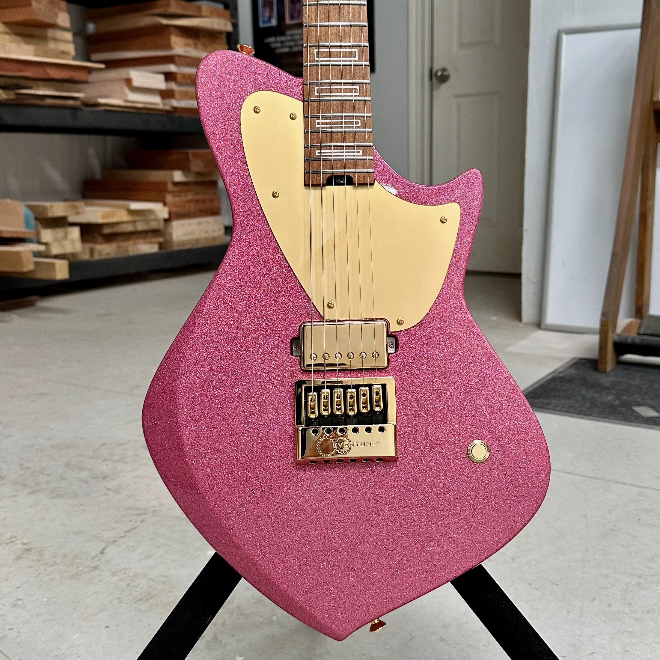 Pink glittery electric Sully guitar with gold hardware, cream-colored pickups, and a wooden neck, standing on a black guitar stand in a workshop or studio.