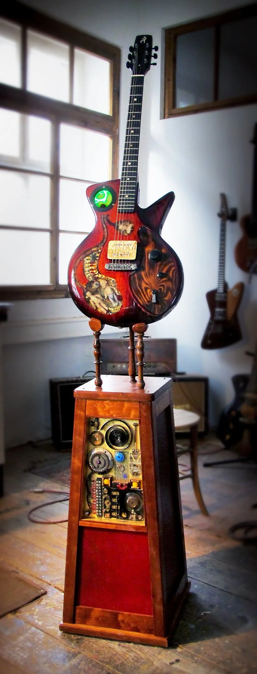 A surreal musical Spalt Instruments sculpture with a guitar body featuring artwork, mounted on a wooden stand on an antique wooden speaker cabinet, inside a room with wooden floors and other guitars on the wall.
