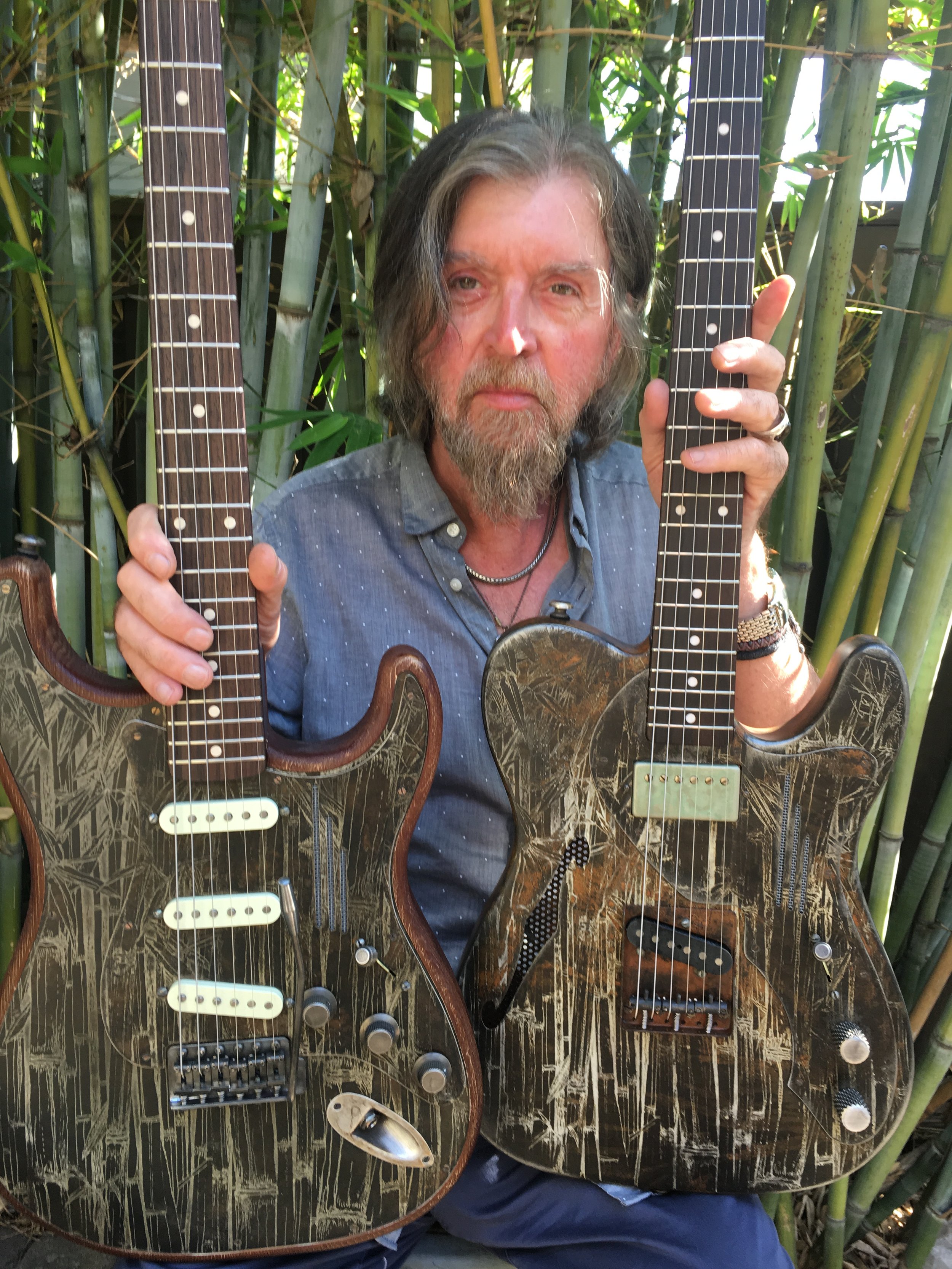 James Trussart with long hair and a beard holding two electric guitars outdoors in front of bamboo plants.