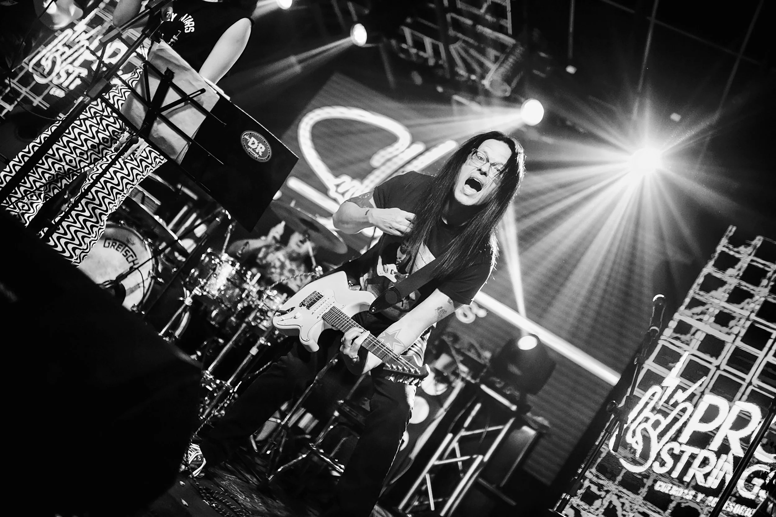 Black and white photograph of a live rock concert with a guitarist in the center, playing an electric guitar and singing passionately. Bright stage lights are visible in the background, along with drums and other band members, and the stage has signage