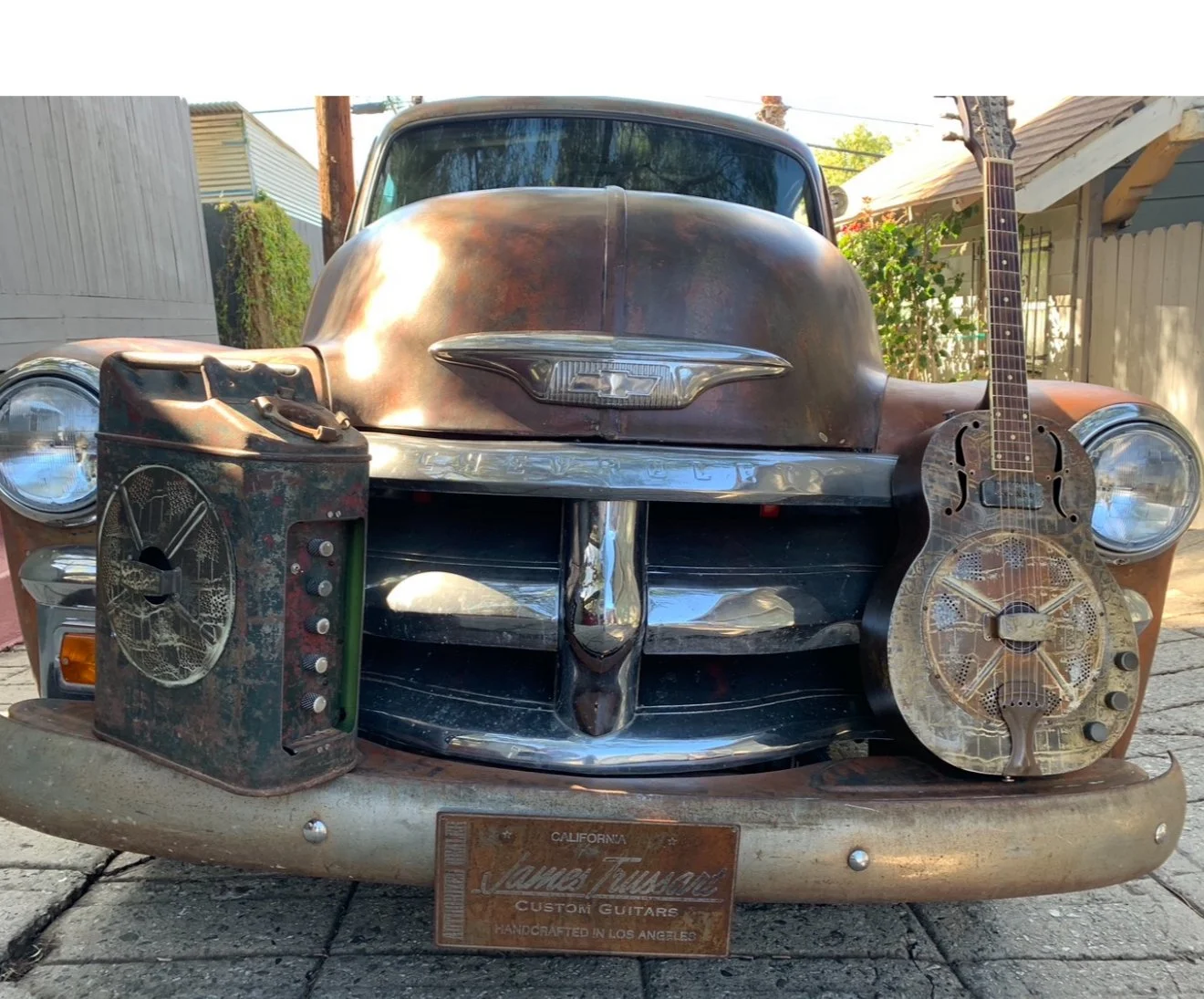 Vintage Chevrolet car with rusted areas on the hood and bumper, and two Trussart guitars placed on the front, one on the left and one on the right, with a rusted metal box on the left. The car has a California license plate.