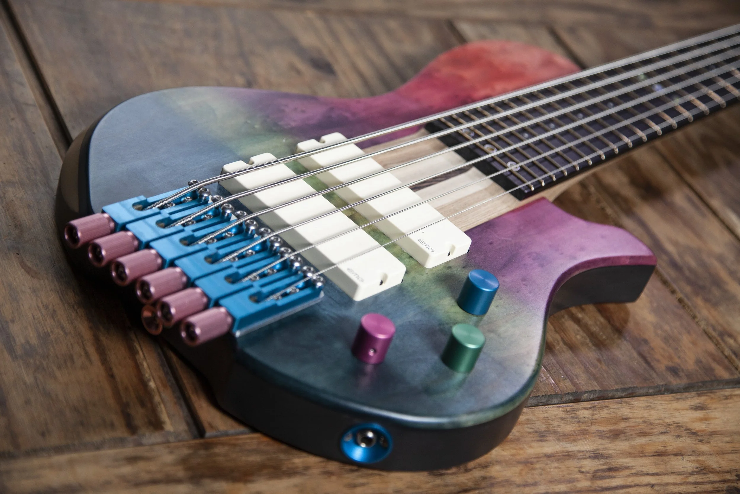 A colorful electric Island Instruments guitar with a rainbow gradient finish, featuring pink and blue tuning pegs, white pickups, and multi-colored knobs, resting on a wooden surface.
