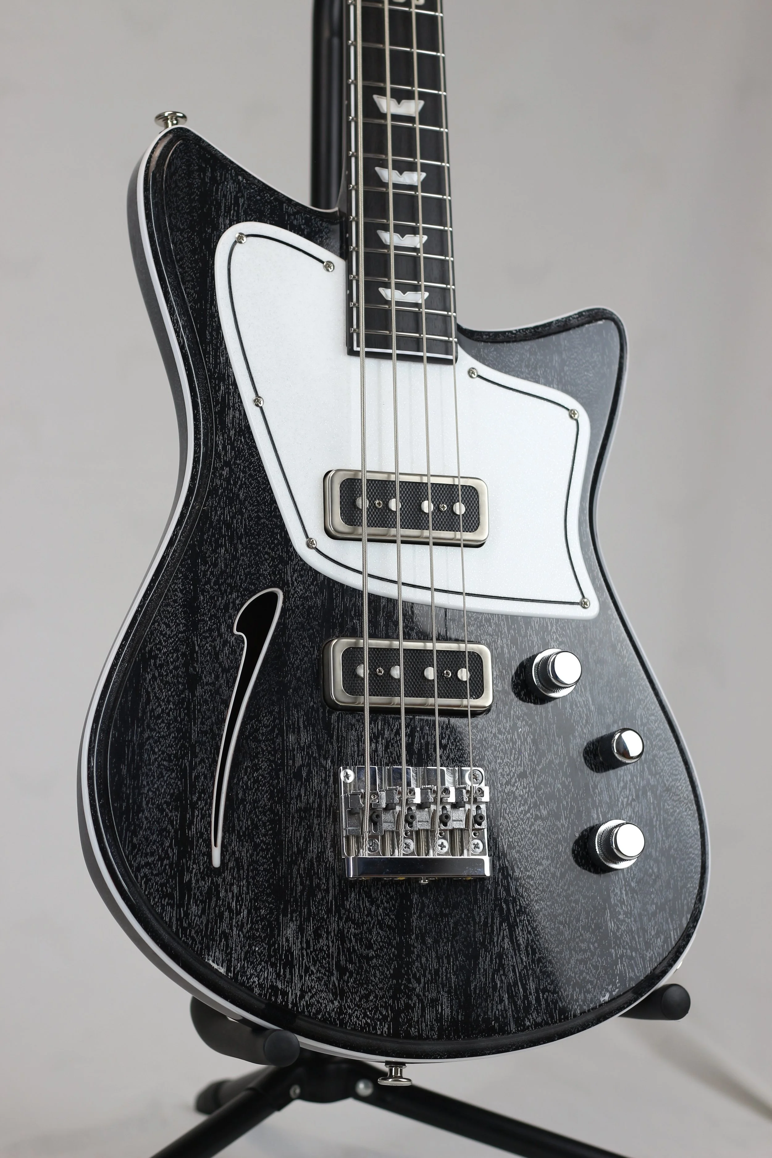 A black electric bass Corsell guitar with two pickups, four control knobs, and a distinctive white pickguard, mounted on a stand against a plain background.