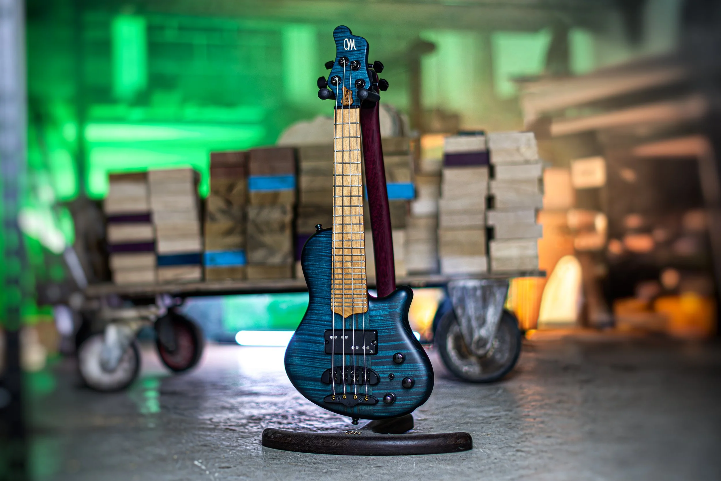 Blue electric Mayones bass guitar with a natural wood neck, standing on a wood stand in a workshop or garage setting, with stacked wooden blocks and a green-lit background.