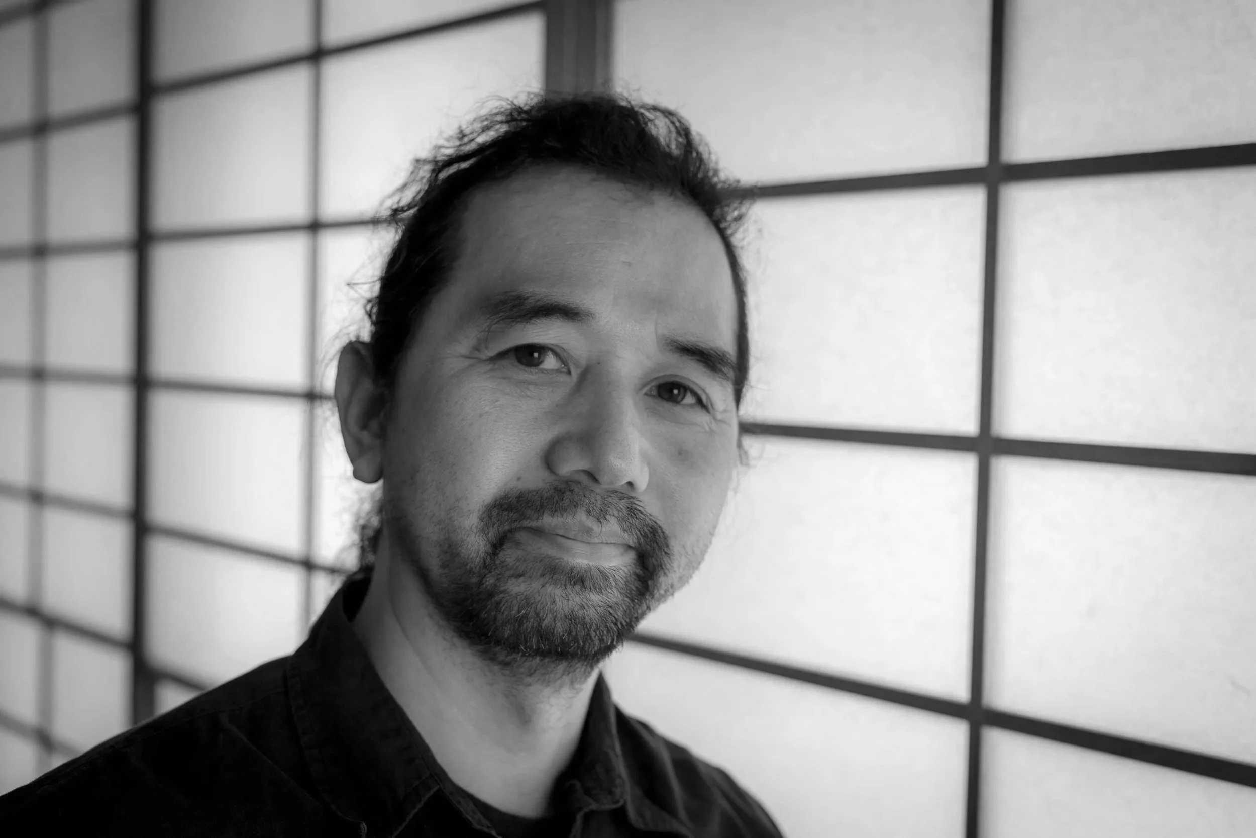 Black and white portrait of Michihiro Matsuda with facial hair, smiling, in front of a grid-patterned wall.