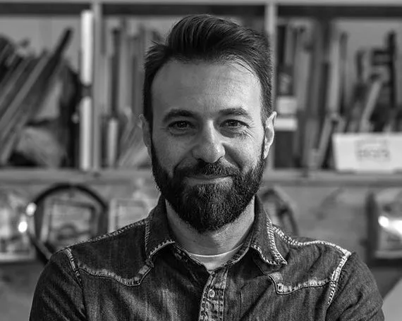 Mirko Borghnio, a man with a beard and dark hair smiling at the camera, sitting in front of a bookshelf filled with books.