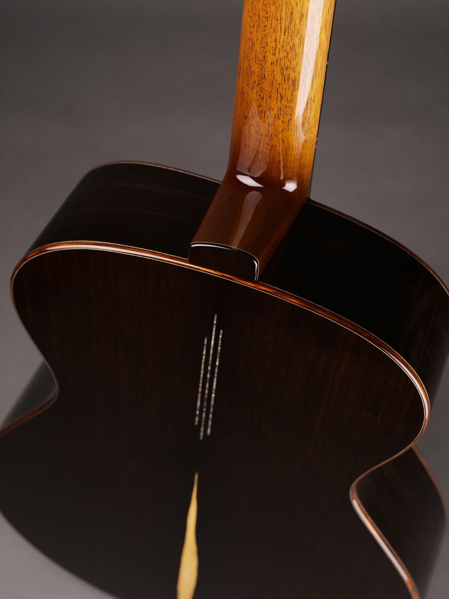 Close-up of the back of an acoustic Isaac Jang guitar showing the dark wood body, neck, and light wood binding, against a dark background.
