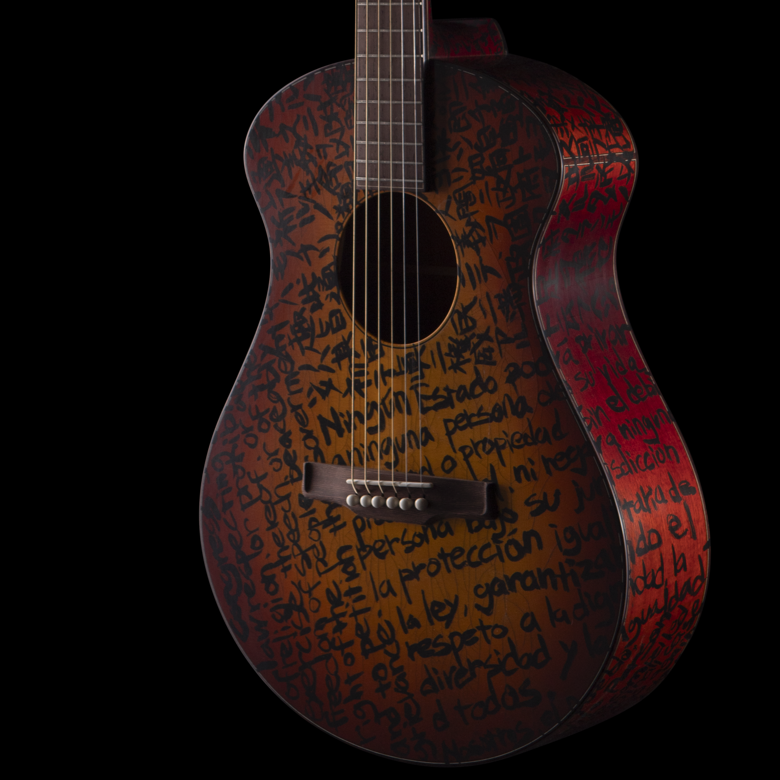 A Matsuda acoustic guitar with black handwritten words covering its body, suspended against a black background.