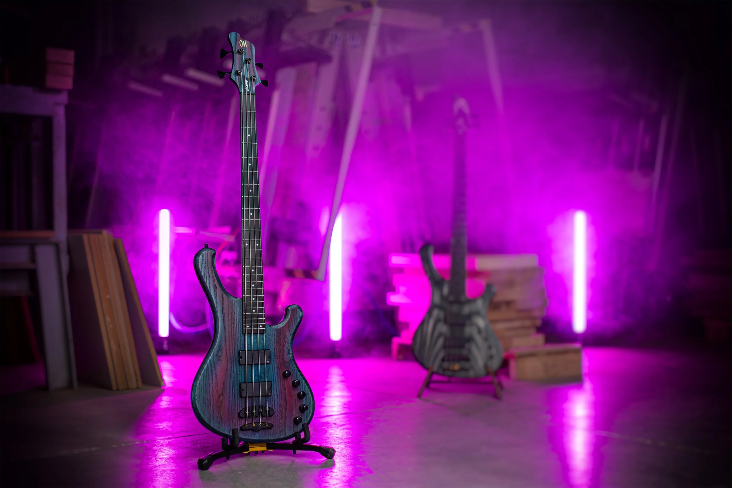 An electric Mayones bass guitar on a stand in a room with a pink-purple smoky fog, with a wall of stacked wooden crates and another electric guitar in the background, illuminated by pink neon lights.