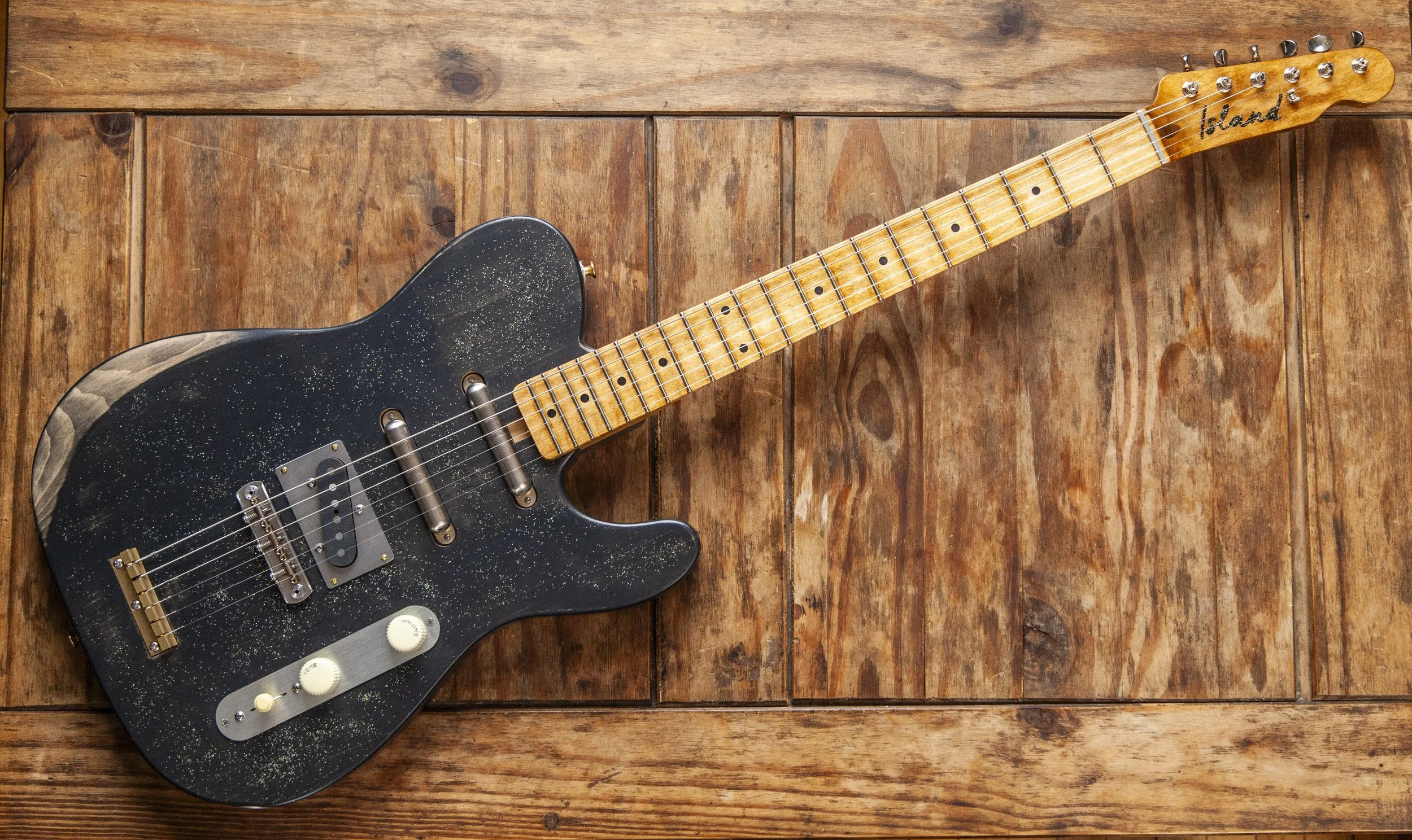 Black electric Island Instruments guitar with a speckled pattern on a wooden surface.